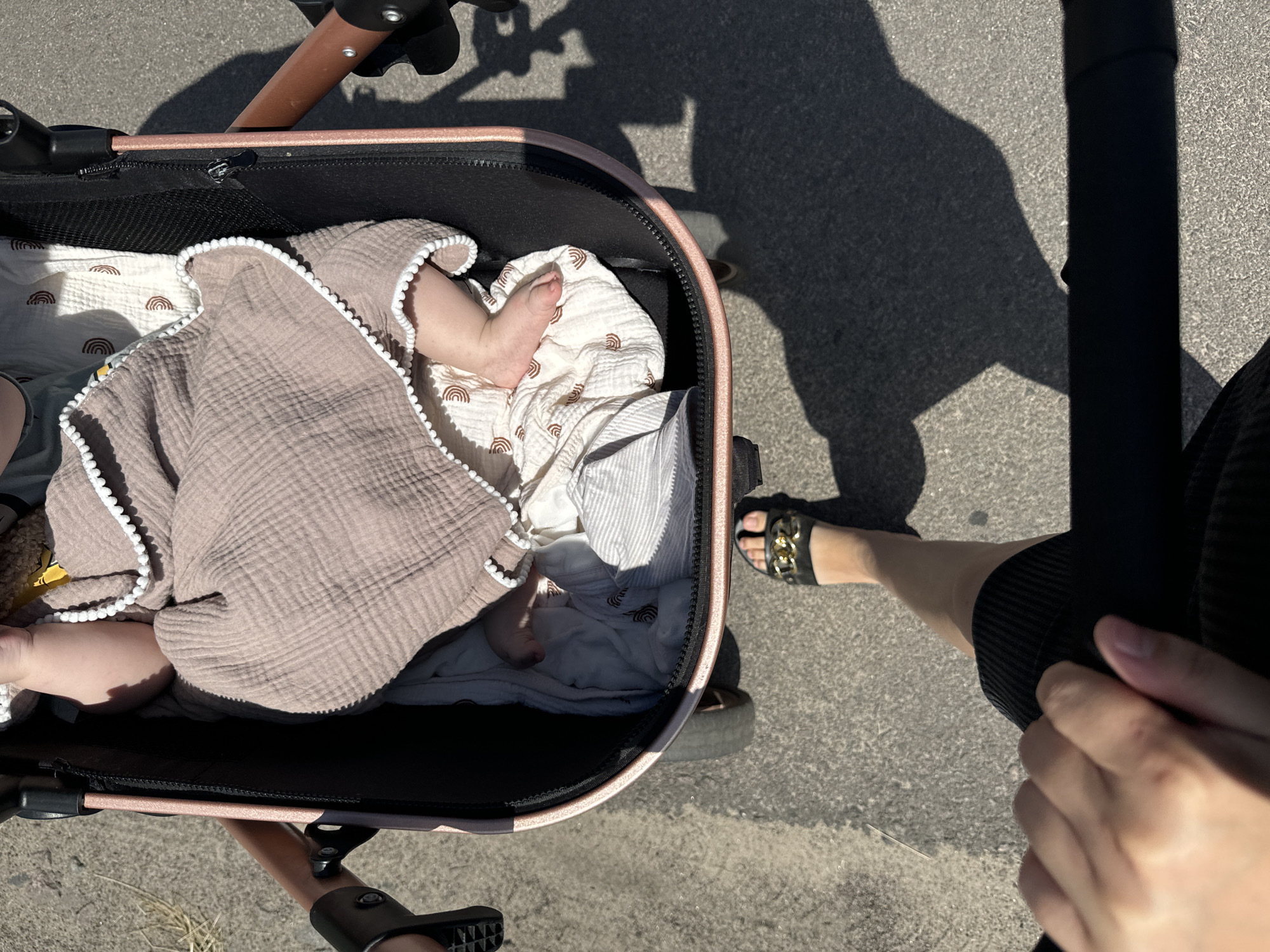 The image shows a close-up view of a baby nestled inside a stroller. The baby is swaddled in a soft, taupe-colored muslin blanket with a white pom-pom trim. A white blanket with a subtle rainbow pattern is visible beneath the baby. The stroller itself has a pinkish-gold metallic frame and a black fabric interior.
In the foreground, a person's legs and feet are visible, indicating they are the one pushing or attending to the stroller. They are wearing black shorts and a pair of black sandals with gold chain embellishments. The perspective suggests the photo was taken from the viewpoint of the person pushing the stroller.
The scene appears to be outdoors, with a sun-dappled asphalt surface suggesting a sunny day. Shadows cast by the stroller and the person are visible on the ground, indicating it is likely daytime. The overall impression is one of a casual outing, possibly a walk in a park or along a street. There is no visible text in the image. The setting is identified as Dovhe, Ukraine.