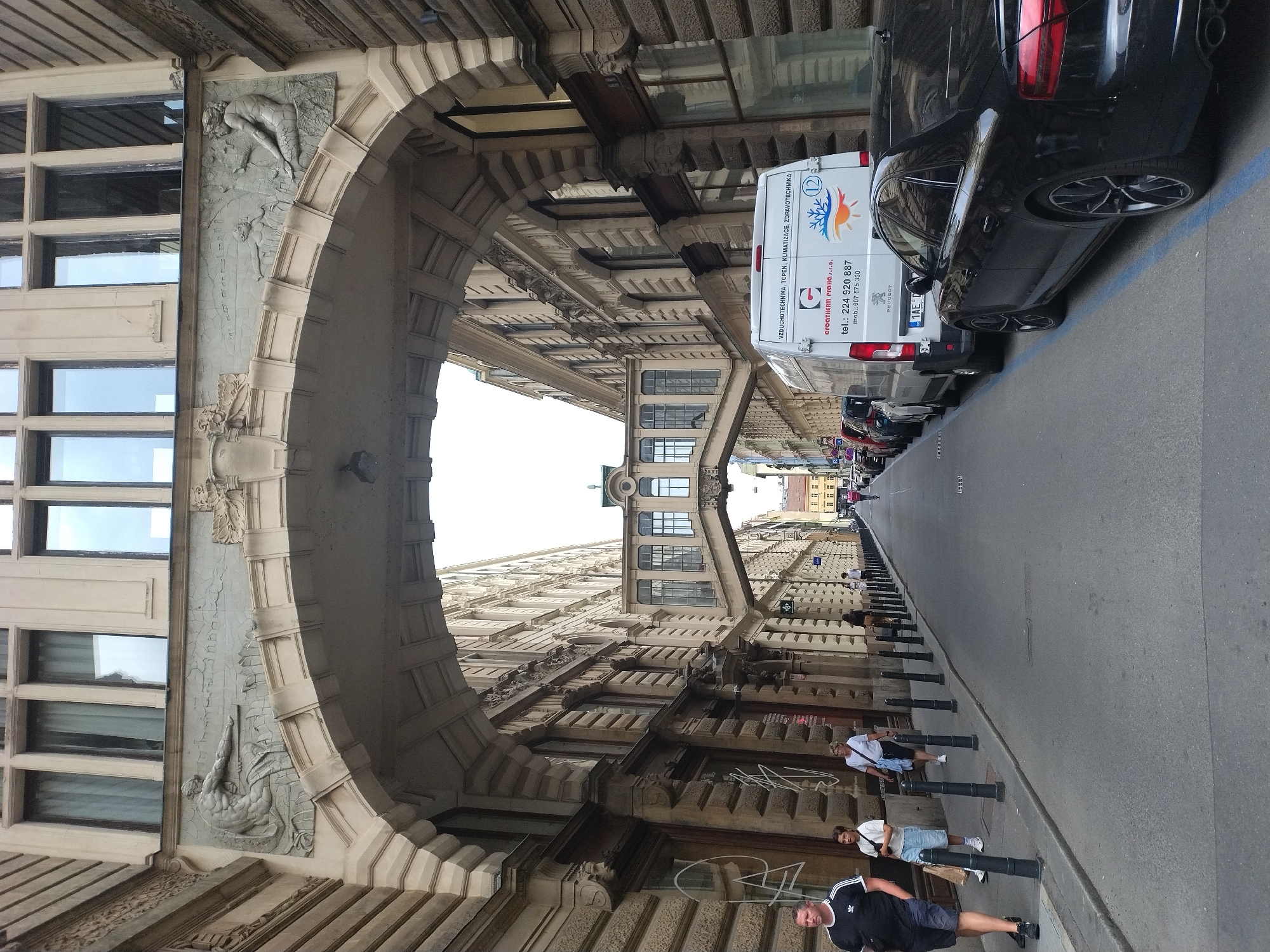 The image depicts a street scene in Prague, Czechia, likely within the Old Town or Lesser Town area, characterized by historical architecture. An imposing stone archway, decorated with relief sculptures of human figures, spans across the street. Beyond the arch, a covered pedestrian bridge connects two buildings at a higher level, creating a distinct architectural feature.
On the left side of the street, several people are walking and standing. A man in a black t-shirt and shorts is walking towards the viewer, while a woman in a white t-shirt and shorts stands near him. Further down the sidewalk, more pedestrians are visible, some walking towards the distance. The sidewalk is lined with black bollards.
On the right side of the street, several vehicles are parked, including a white van with company logos and contact information ("VZDÚCHOTECHNIkA, TOPENí, KLIMATIZACE, ZDRAVOTNICKÉ") and a black car with its rear facing the viewer. The street itself is paved with asphalt and appears to be a one-way street, with a blue line painted on the road.
The buildings lining the street are multi-story structures with traditional facades, featuring numerous windows and decorative architectural elements. The overall impression is of a historic European city street, with a mix of old and new elements. The sky is overcast.