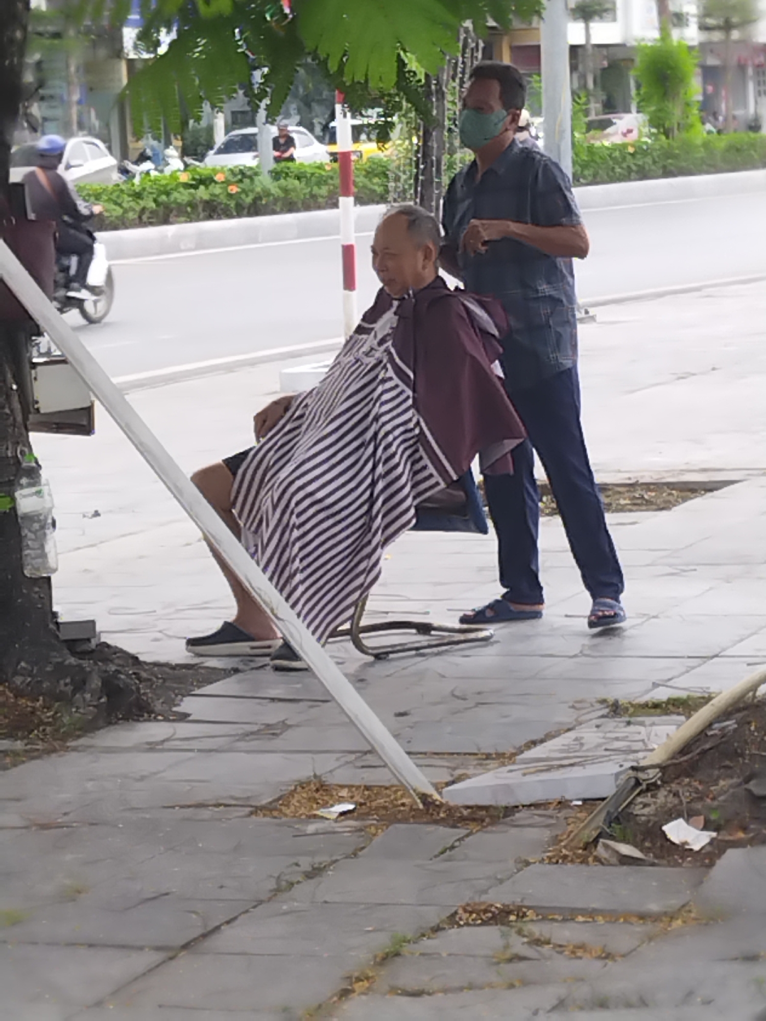 The image depicts an outdoor street barber service in Hong Gai Ward, Vietnam, during daylight hours. Two men are the central subjects. An older man, the customer, sits in a metal barber chair on a paved sidewalk, draped in a distinctive striped white and dark grey barber cape over a maroon outer cape. He appears relaxed, looking towards his left with a neutral expression.
Behind him stands a younger man, identified as the barber, wearing a green face mask, a dark blue plaid shirt, and dark blue trousers, along with blue sandals. His hands are positioned near the customer's head, suggesting he is actively giving a haircut or preparing to do so.
The setting is a vibrant urban street. A wide road with traffic flows in the background, where a person on a white scooter and several cars can be seen. A green median strip with bushes and trees separates the traffic lanes. On the sidewalk, a large tree provides shade, with a plastic water bottle hanging from its trunk. The grey tiled sidewalk shows some scattered leaves and debris, and a red and white striped pole stands near the curb. The overall atmosphere suggests a normal, busy day in a Vietnamese city. No visible text is clearly discernible.