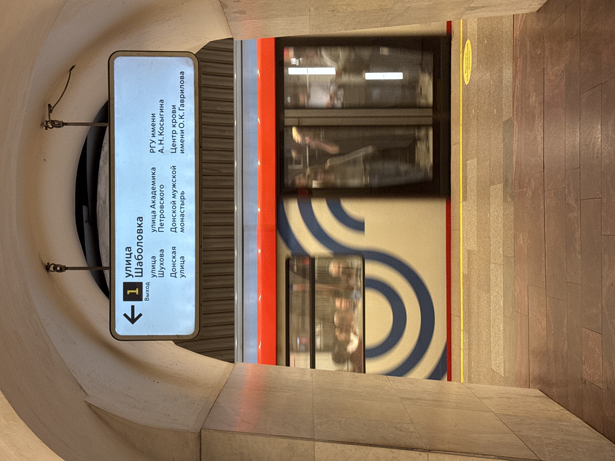 The image depicts a dynamic scene within a Moscow Metro station, viewed from an arched passageway that opens onto a platform. A subway train, characterized by its red, white, and blue livery and a distinctive blue wave pattern on its side, is captured in motion, creating a blur that conveys its speed. Through the train's windows, blurred figures of passengers are discernible.
Hanging prominently from the archway on the left, a bright, backlit sign provides information in Cyrillic. The sign indicates "1 улица Шаболовка" (Shabolovka Street), likely identifying the station or a primary nearby location. An arrow points left, accompanied by "Выход" (Exit). The sign further lists several destinations: "улица Шухова" (Shukhova Street), "улица Академика Петровского" (Academician Petrovsky Street), "Донская улица" (Donskaya Street), and "Донской мужской монастырь" (Donskoy Monastery). Additionally, it points towards "РГУ имени А. Н. Косыгина" (Russian State University named after A. N. Kosygin) and "Центр крови имени О. К. Гаврилова" (O. K. Gavrilov Blood Center).
The platform floor is composed of beige and brown tiles, demarcated by a vibrant yellow safety line that runs parallel to the tracks. A partially visible yellow circular sticker with text is also on the floor. The setting is artificially lit, characteristic of an underground transit system, and evokes the busy atmosphere of the Moscow Metro.