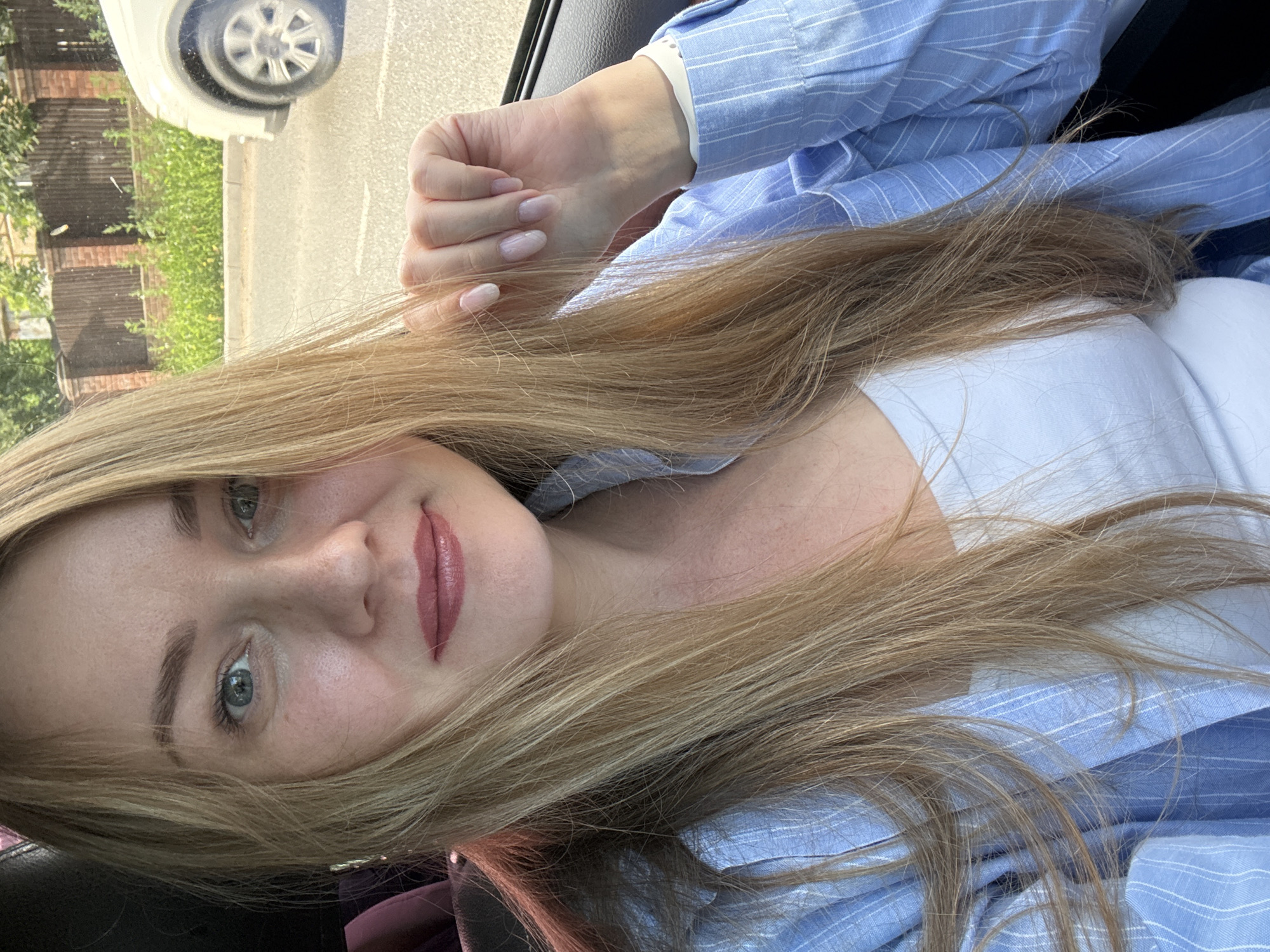 A medium shot captures a Caucasian woman with long blonde hair and fair skin sitting in a car, presumably in Chaikovsky, Russia. She is wearing a light blue striped button-up shirt over a white top and has a light application of makeup. Her left hand rests on her lap, showing manicured nails with a pale pink hue. In the background, a blurred view through the car window reveals a sidewalk, greenery, a wooden fence, and a portion of a white car. The lighting suggests it is daytime.