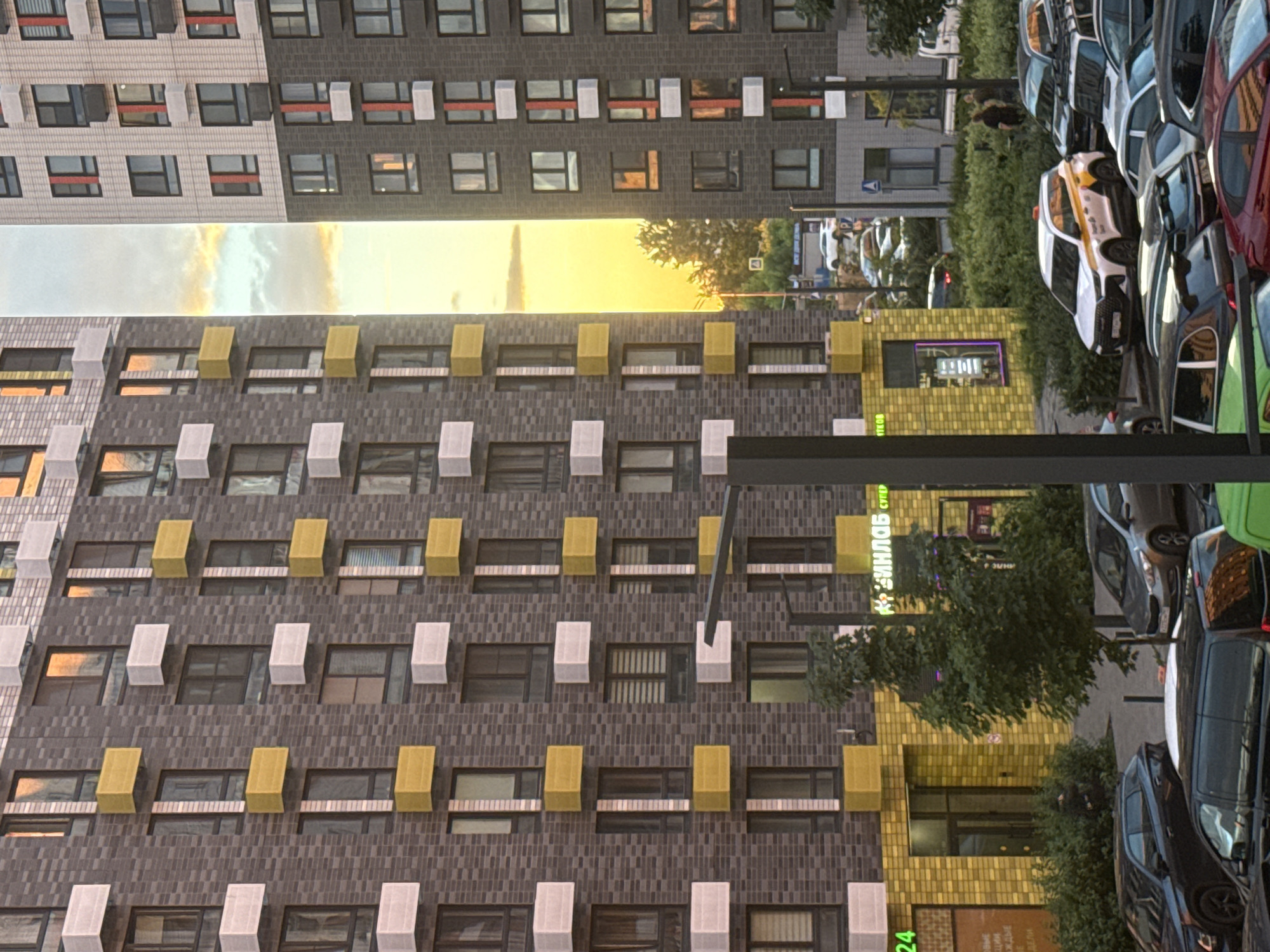The media file displays a street scene in Drozhzhino, Russia, characterized by modern apartment buildings and a variety of parked cars. The perspective is somewhat distorted, giving the impression of a tall, multi-story structure filling the frame. Several cars are visible, including a distinctive white and yellow car that appears to be a taxi, with visible text on its side. Other vehicles include a red car, a green car, and several darker colored cars, some of which are partially obscured.
The apartment buildings feature a patterned brick facade with contrasting architectural elements such as white and yellow rectangular protrusions above some windows. The windows themselves are dark, reflecting the surroundings. A bright, sunlit sky with scattered clouds suggests it is daytime, likely late afternoon or early evening given the warm hues of the sunlight. There are also areas of dense green foliage, indicating the presence of trees and bushes, possibly in a park or courtyard area adjacent to the buildings and parking lot.
On one of the buildings, a yellow storefront with a glass entrance is visible. Above the entrance, illuminated green text in Cyrillic characters can be seen. Some of this text appears to read "ЦИФРЛАВ СУПЕР". Another sign with green text is partially visible near the bottom of the frame, with the number "24" prominent. The overall scene depicts an urban residential environment with elements of everyday life, including transportation and commercial activity.