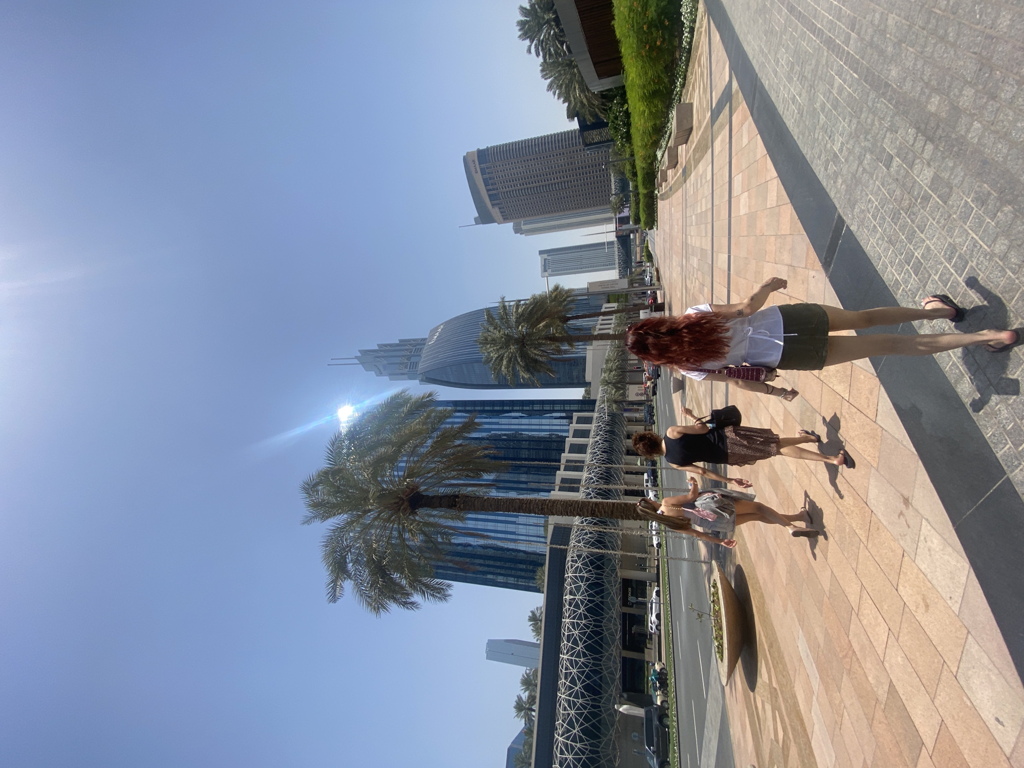 The image depicts a daytime scene in Dubai, United Arab Emirates, with a group of three women walking along a paved walkway. The setting includes a cityscape with skyscrapers and palm trees, under a clear blue sky. The sun is visible, creating a bright glare. The women are casually dressed, with the closest one wearing a white shirt and olive shorts. The other two women are wearing dark clothing, and walking in the direction of the camera. The architecture is modern and urban, with various buildings and structural elements in the background. There are also cars parked on the side of the street.