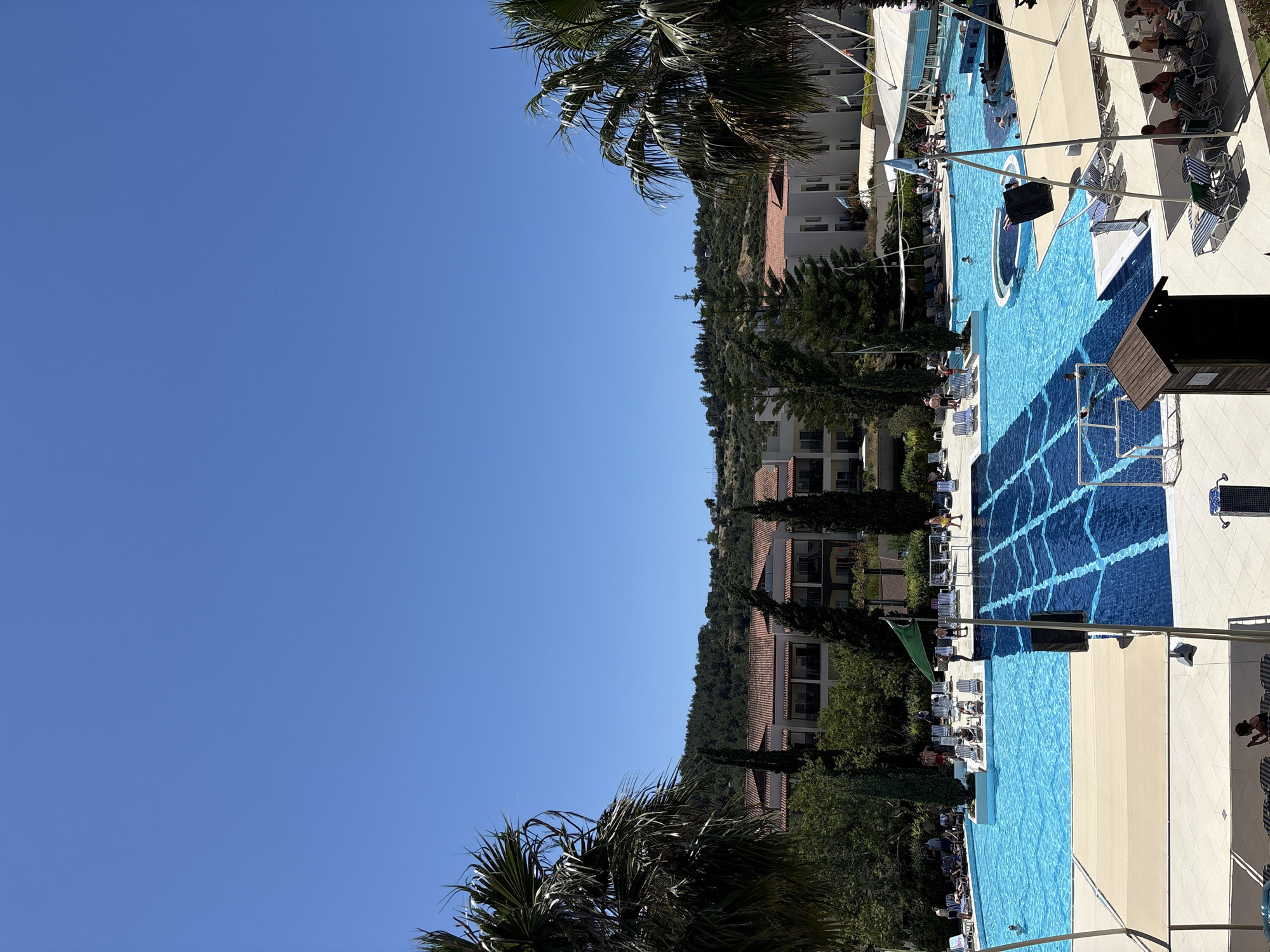 The image shows a partially filled swimming pool at a resort or hotel in Selçuk, Turkey, with clear blue water. Some people are swimming, and others are sunbathing on lounge chairs lining the pool. The pool area is well-maintained with beige and white paving. There are hotel buildings, trees, and clear blue sky in the background. Some palm trees are visible on the right and left of the image.