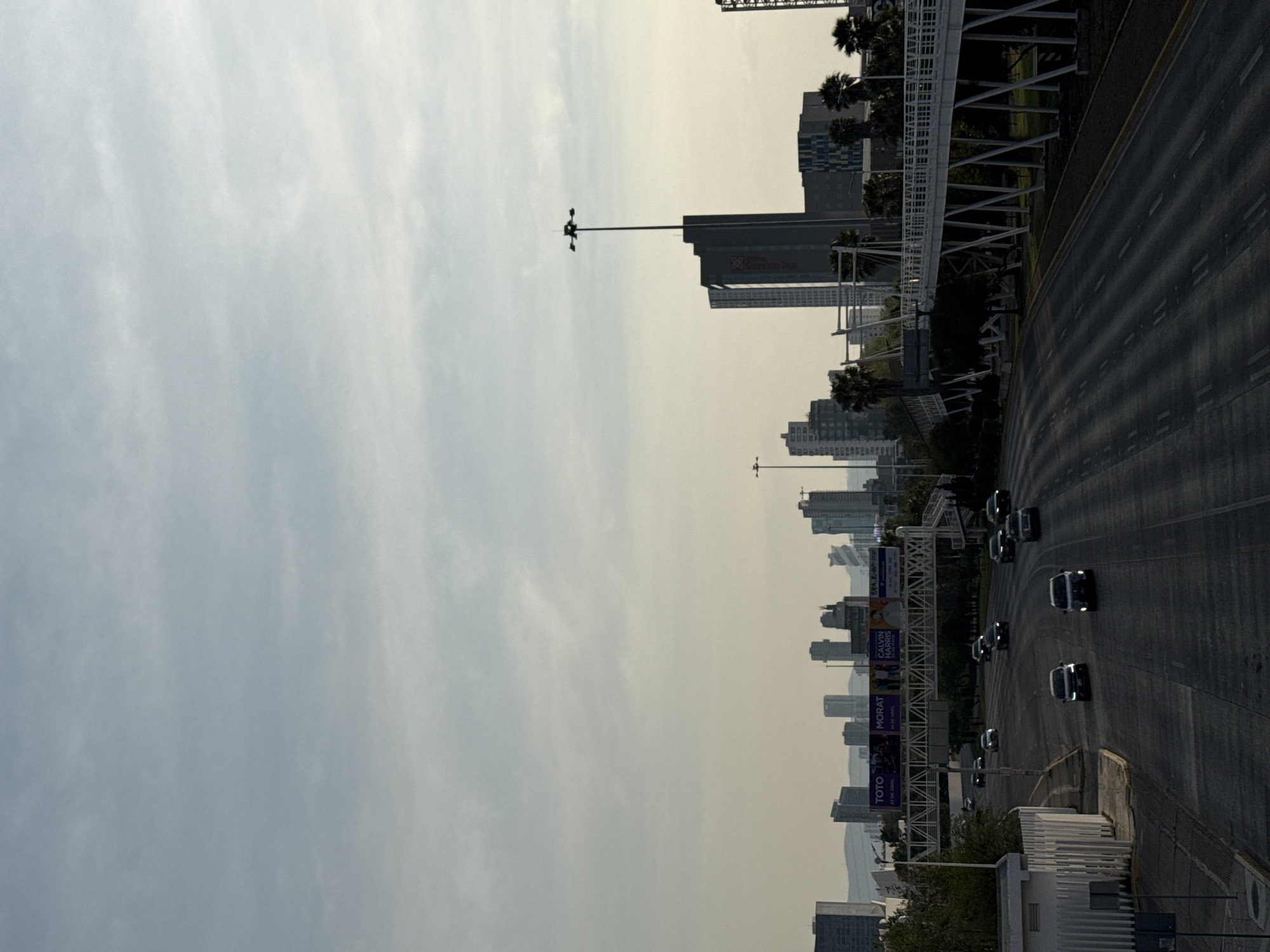 This image shows a street scene in Puebla, Mexico, with tall buildings lining the road. The sky is overcast. Several cars are driving on the road. A pedestrian bridge crosses the street. Banners and signs are visible on the bridge.