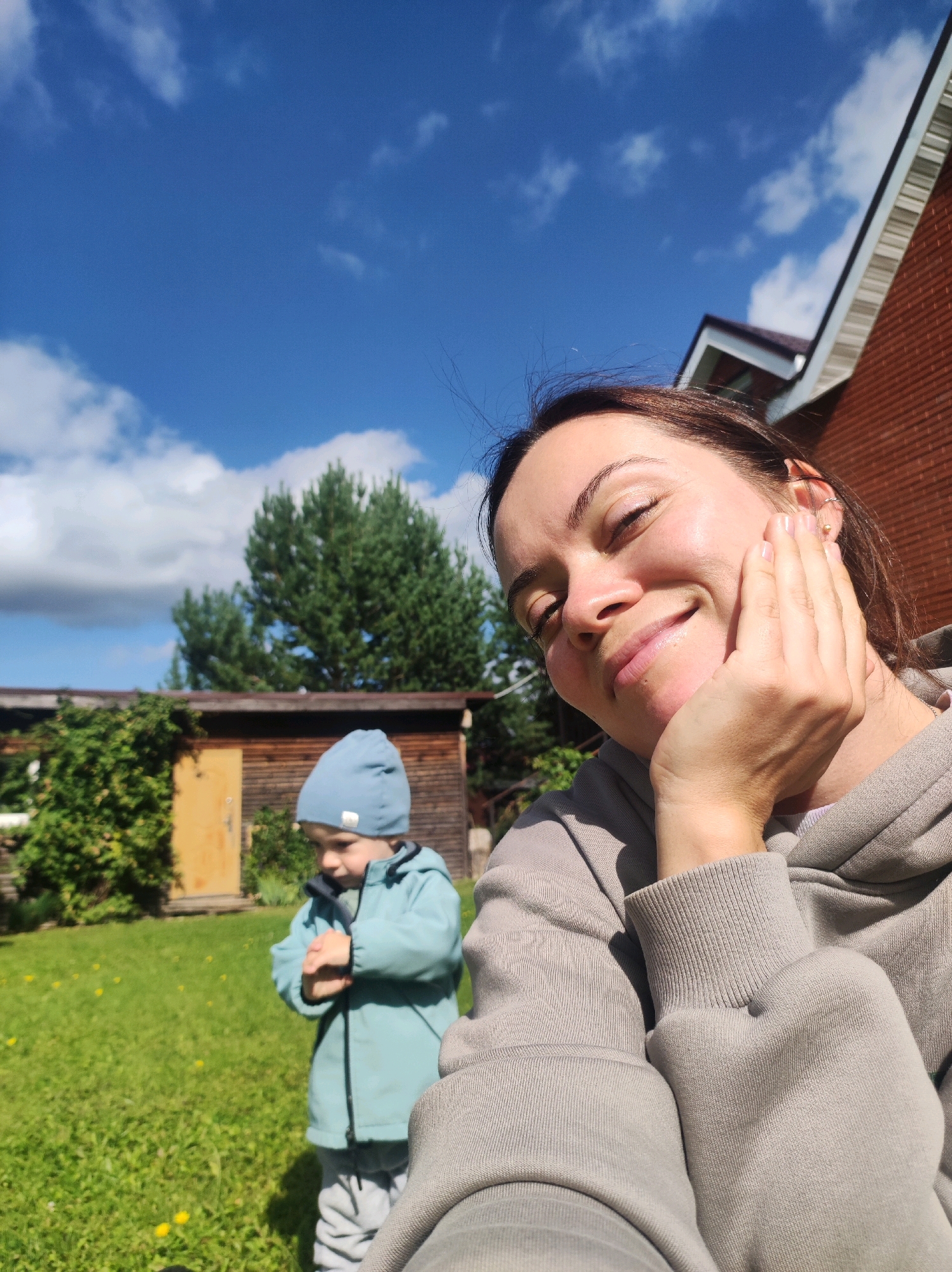 In an outdoor setting, a woman is captured in a close-up selfie with a child in the background. The woman, with her eyes closed and a smile, rests her hand on her cheek. She is wearing a grey hooded sweatshirt. Behind her, a young child in a blue hat and jacket stands with arms crossed, looking away. The environment includes a grassy lawn with scattered yellow flowers, a wooden shed with a yellow door, and trees. A brick house with a white trim roof is visible to the right. The sky is blue with scattered clouds. The location is identified as Фоки, Russia.