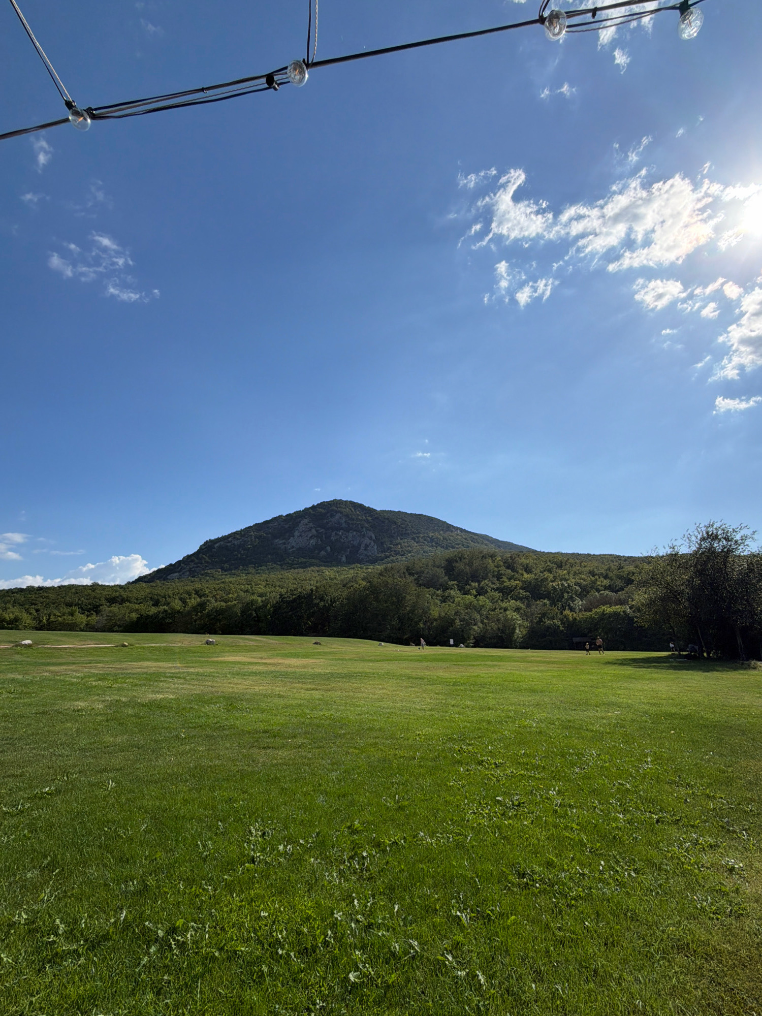 The image depicts a wide, lush green meadow with a large, tree-covered mountain dominating the background. The sky is a clear, vibrant blue with scattered white clouds, and the sun is visible in the upper right corner, casting a bright glare. Several small, indistinct figures are scattered across the meadow, suggesting people are enjoying the outdoors. Some appear to be walking or standing, while others might be sitting. In the foreground, a string of decorative lights is strung across the top of the frame, suggesting this is an outdoor gathering space or event area. The overall impression is one of a peaceful, scenic natural landscape on a sunny day.