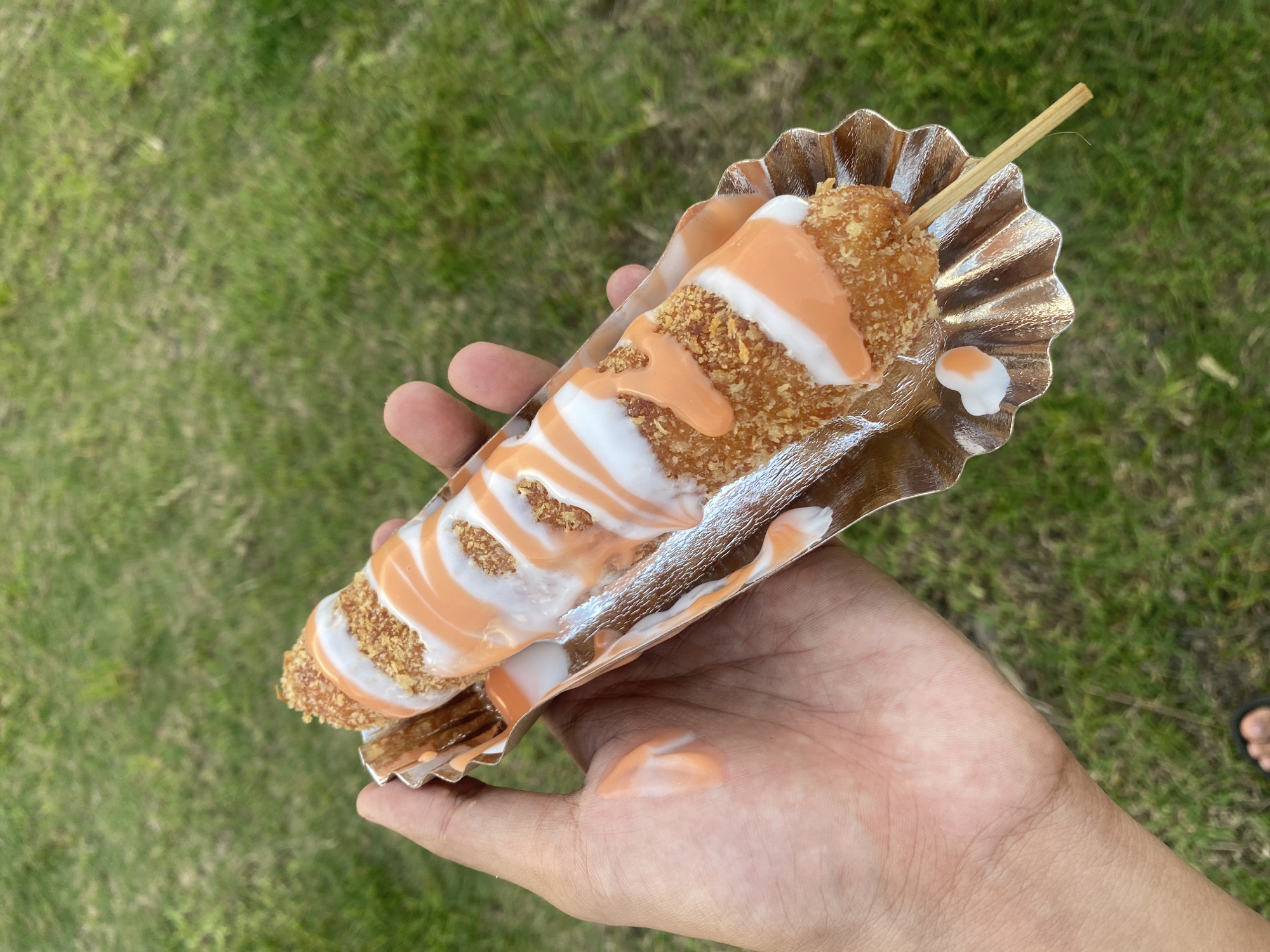 A close-up, high-angle shot captures a person's hand holding a street food item against a blurred green grass background, suggesting an outdoor setting like a park or field in Davao City, Philippines. The food is a long, fried, golden-brown snack, possibly a corndog or similar breaded item, served on a wooden stick. It is heavily drizzled with contrasting white and orange sauces, creating a striped pattern across its surface. The fried coating appears crunchy, with visible breading or crumbs.
The snack rests in a fluted, metallic-colored paper tray or boat, which the hand is holding from underneath. Some of the orange and white sauce has dripped onto the palm of the hand, indicating recent consumption or handling. The lighting suggests it is daytime. In the background, a portion of a bare foot wearing a dark sandal or slipper is faintly visible, further confirming a casual, outdoor environment. There is no visible text in the image.