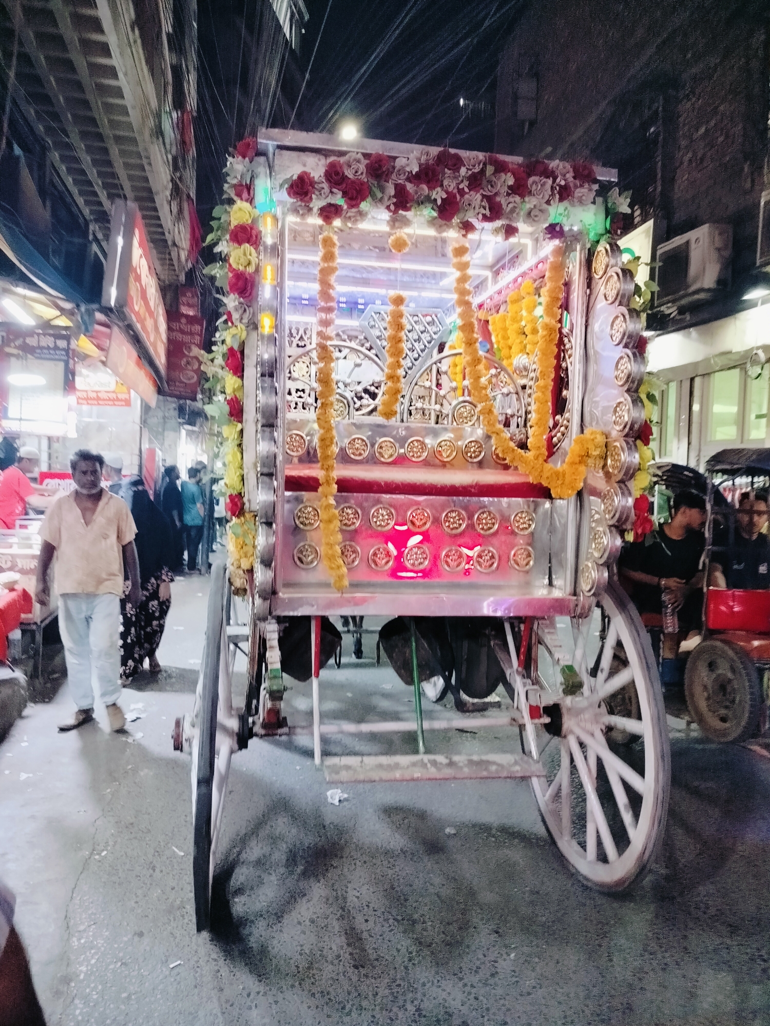 In a bustling street scene in Dhaka, Bangladesh, a decorated carriage, likely a rickshaw or a ceremonial vehicle, takes center stage. Adorned with vibrant red and white flowers and yellow garlands, the carriage features intricate metalwork and illuminated panels. The street is narrow, with overhead wires and buildings lining either side, suggesting a commercial or residential area.
In the foreground, a man in a light-colored shirt and pants walks along the wet, uneven pavement. Behind him, a woman in a patterned skirt and headscarf is visible, along with other pedestrians and a glimpse of street vendors. To the right, a rickshaw driver and passengers are seated in another vehicle, partially obscured. The overall impression is one of a lively urban environment with a blend of traditional and modern elements, common in Dhaka. The lighting suggests it is either dusk or night, with artificial lights illuminating the scene.