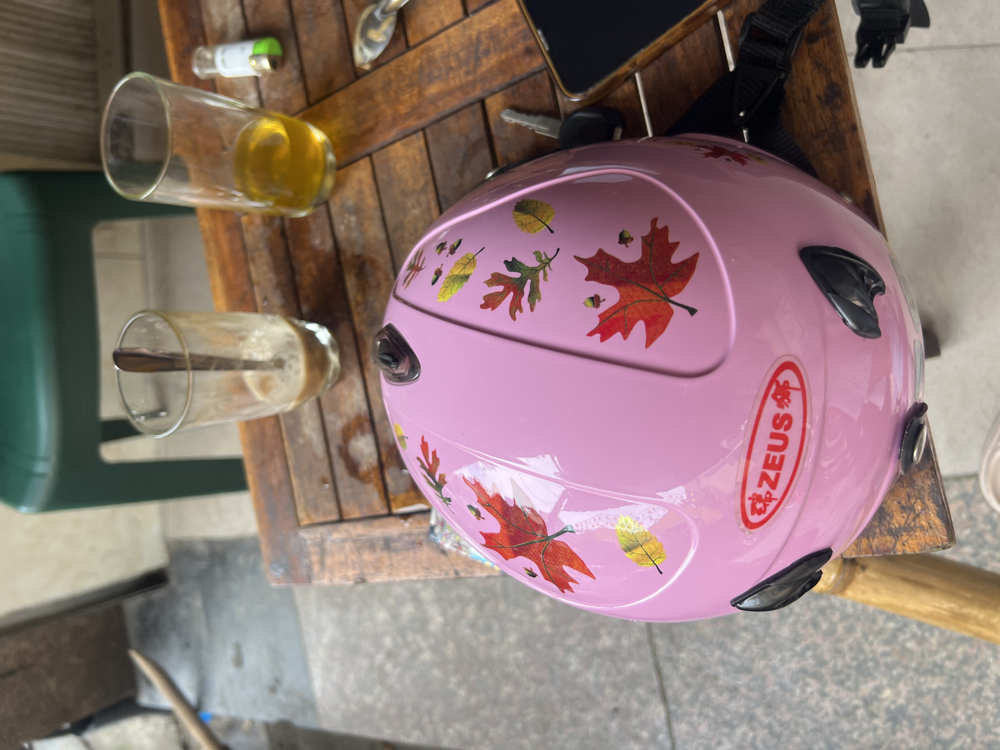 In a cafe setting in Hanoi, Vietnam, a pink helmet adorned with autumn leaf decals rests on a rustic wooden table. Two glasses, one containing a milky coffee and the other a golden liquid, sit nearby, alongside a small lighter and car keys. A smartphone is also visible on the table. The background shows a glimpse of a green chair and a concrete floor, suggesting an outdoor or semi-outdoor seating area.