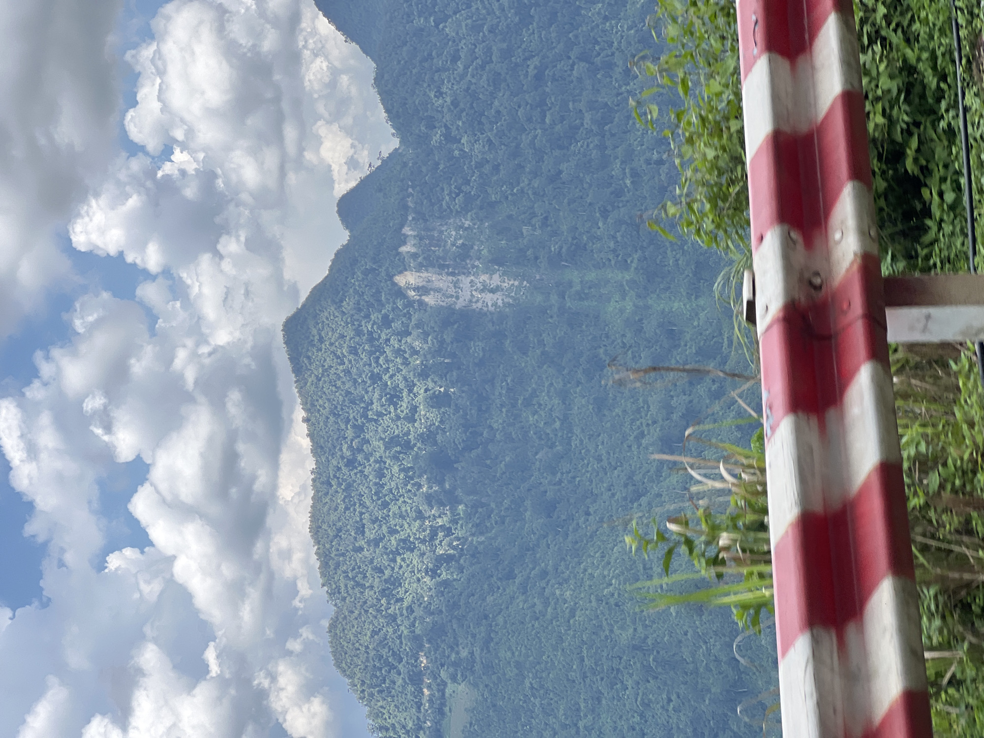 A roadside guardrail with red and white stripes is in the foreground. Beyond the guardrail, lush green mountains rise under a partly cloudy sky. A pale, rocky scar is visible on the face of one of the mountains. The vegetation is dense, indicating a tropical or subtropical climate. The scene appears to be a scenic overlook or a roadside view in a mountainous region. The image is oriented vertically. There are no people or animals visible. The location is Minh Tân Commune, Vietnam.