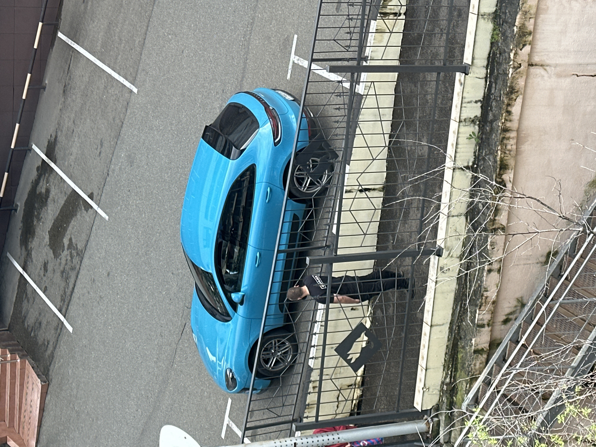 The image depicts a light blue Porsche parked in a marked parking space in Sochi, Russia. A man wearing a black shirt and pants is lying on a fenced-in area next to the car. The parking area is adjacent to a weathered concrete structure with a metal fence. Bare tree branches are visible in the foreground. The photograph appears to be taken from an elevated perspective, looking down on the scene.