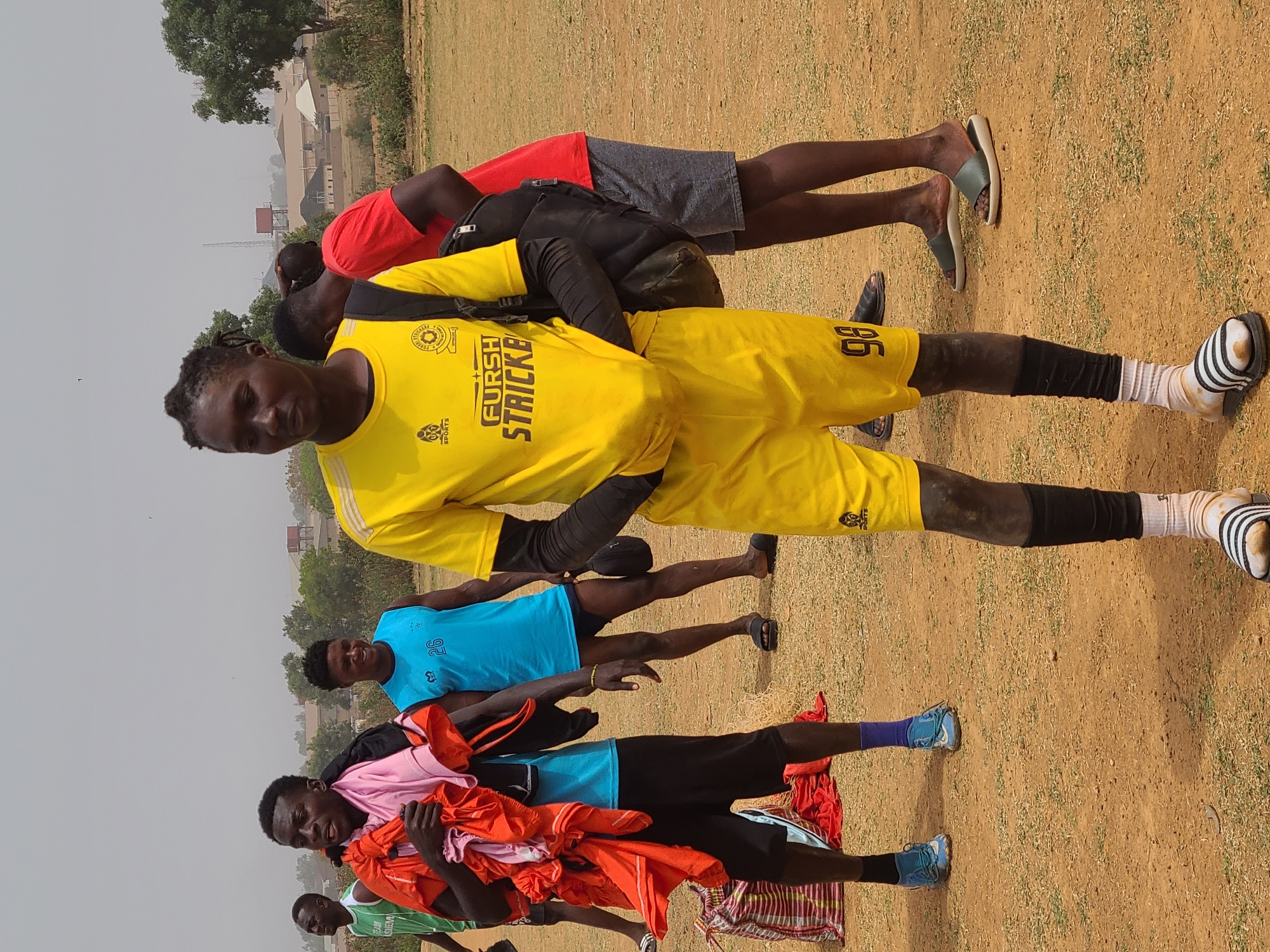 A group of men stand on a grassy, dirt field. In the foreground, a man wearing a yellow jersey and shorts with the number 96 is looking at the camera. He has black sleeves on his arms and a black backpack. Behind him, a man in a red shirt and grey shorts stands with his back to the camera. Further back, another man wears a blue shirt. In the background, other men stand with colorful clothing items draped over them. The ground is a mix of dirt and patchy grass. In the distance, buildings and trees are visible under a hazy sky. The location is Barikin Jirga Summa, Nigeria.