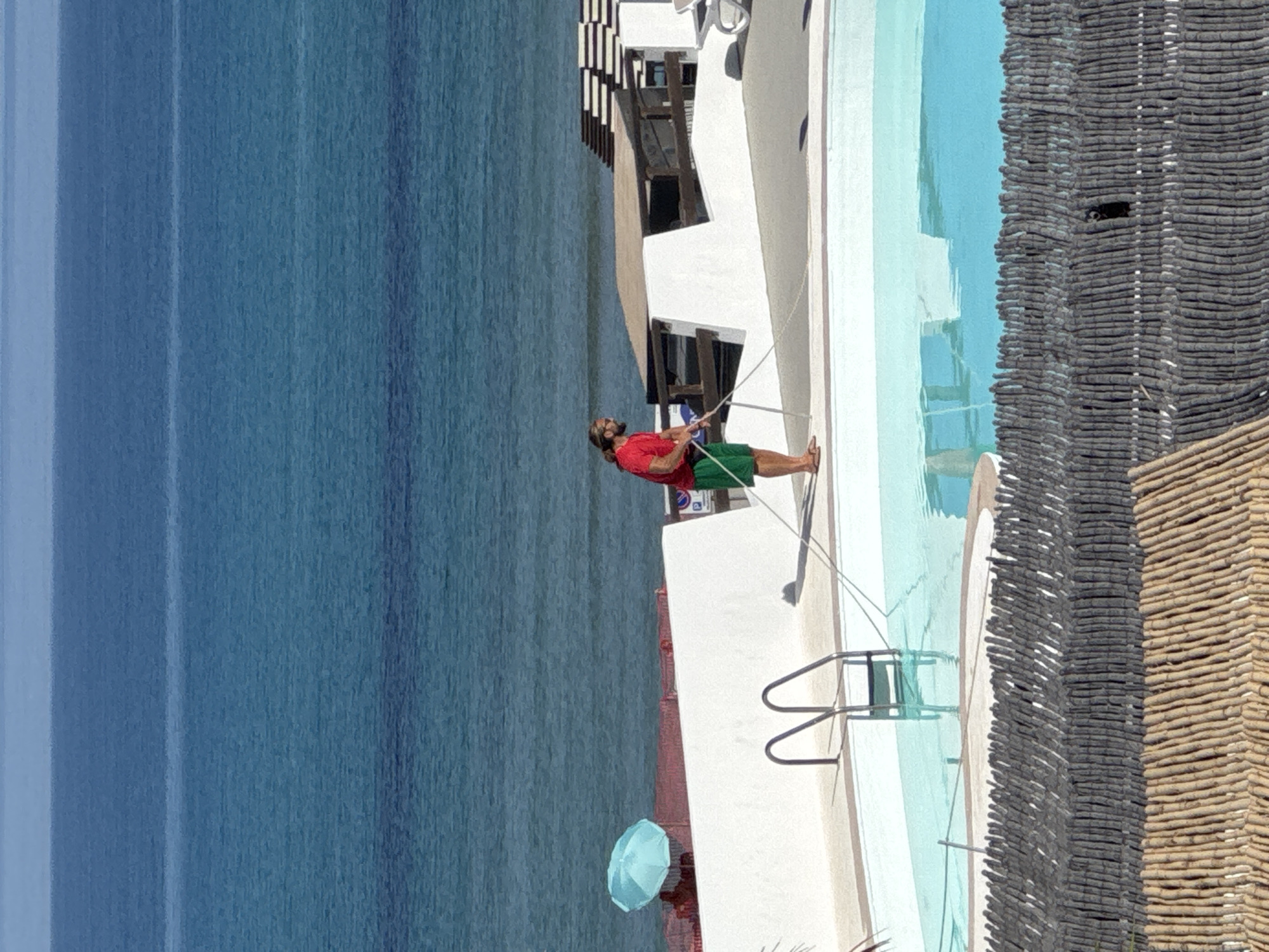 The image captures an outdoor coastal scene identified as being in Isola delle Femmine, Italy, on a bright, clear day. The upper two-thirds of the frame features a calm, blue-green sea, displaying subtle color gradations indicative of varying depths. A distinct horizon line separates the sea from a pale blue sky.
In the mid-ground, a white, angular structure, resembling a low building or retaining wall, spans the width of the frame, separating the pool area from the ocean. Behind this wall, to the left, a light blue beach umbrella is partially visible, alongside red structures that could be lounge chairs or barriers. To the right, a multi-tiered wooden deck structure with parallel dark and light bands is present, next to a white lounge chair. A partial "P" sign, possibly for parking, is visible on the white wall near the umbrella.
A male figure stands on the edge of a light blue, irregularly shaped swimming pool in the mid-to-foreground. He has long brown hair and a beard, and is dressed in a red short-sleeved shirt, green shorts, and light-colored sandals. He holds a long white pole with both hands, extending it into the pool, indicating he is performing maintenance. Two metal ladders with curved handrails are submerged in the pool on the left side.
The immediate foreground reveals two distinct layers of fencing. The upper fence is constructed from numerous closely packed, thin, dark grey or black vertical wooden sticks. Beneath it, the lower fence is composed of similar, light brown or natural-colored bamboo sticks, also arranged vertically and densely packed.