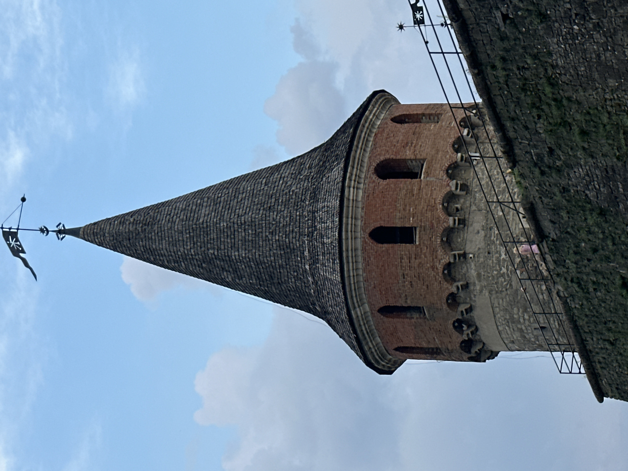 The image shows the Kamianets-Podilskyi Castle in Kamianets-Podilskyi, Ukraine. The castle is a historic fortress located on a rocky island, surrounded by the Smotrych River. The image focuses on a circular tower with a conical roof, made of brick and stone. A person in a pink dress is visible on a walkway leading up to the tower. The sky is cloudy and blue, with a weather vane at the top of the tower.