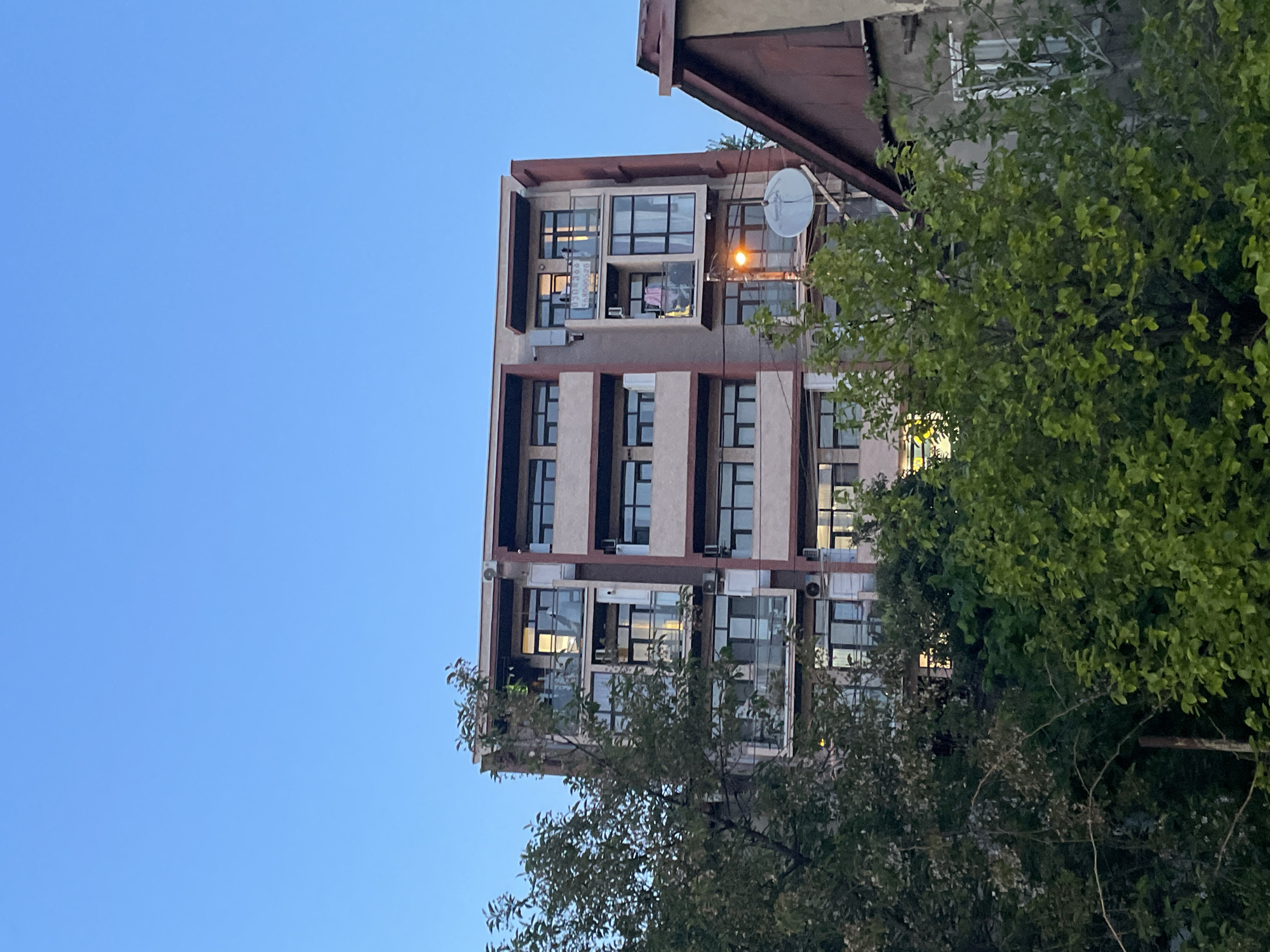 This image depicts a multi-story building, likely residential or mixed-use, viewed from a low angle against a clear, light blue sky. The building features a light-colored facade with darker vertical bands and numerous large windows. Several enclosed balconies or extensions are visible, along with multiple air conditioning units mounted on the exterior. A satellite dish is attached to the upper right side of the building. An older, adjacent structure with a dark, sloped roof is partially visible to the right.
Lush green trees are prominent in the foreground and mid-ground, partially obscuring the lower and right portions of the building. The clear blue sky suggests fair weather. Illuminated interior lights within some windows indicate that the time of day is likely dusk or early evening.
A significant detail is a vertical sign or banner hanging from a window on one of the upper floors. This sign contains text in Georgian script, which reads "გასაქირავებელია" (Gasak'iravabelia), translating to "For Rent," followed by a phone number, "599059520". No specific human activity is explicitly visible, but the illuminated windows suggest occupancy. The scene portrays an urban setting.