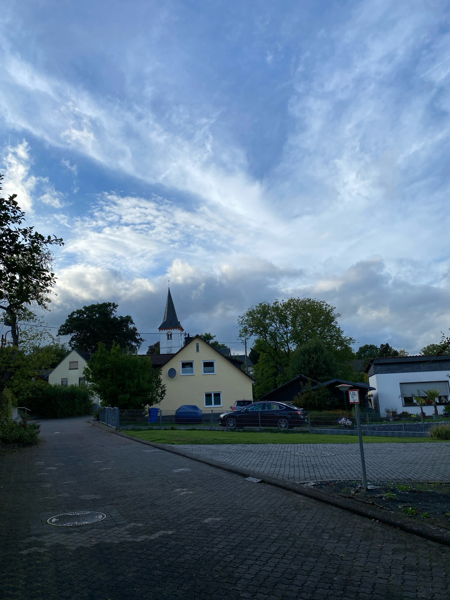The image shows a street scene in the city of Flammersfeld, Germany. The sky is cloudy with patches of blue visible. In the background, a church steeple with a cross on top is visible, surrounded by trees. Several houses are also present, with a yellow house being prominent in the center. A dark-colored car is parked in front of one of the houses. The street in the foreground is paved with interlocking bricks. A traffic sign indicating "H 4,80 0,1 0" is visible near a fence. The overall scene depicts a quiet residential area.