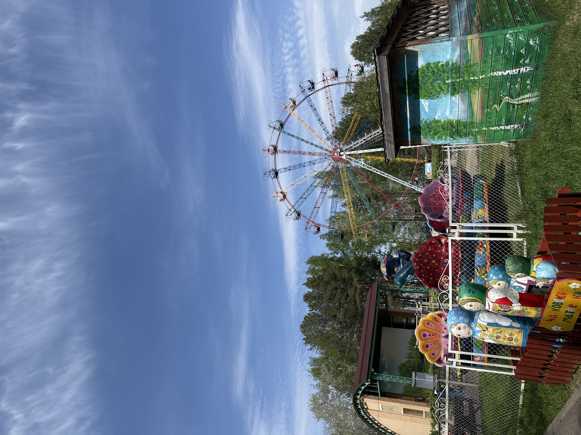 The image shows an amusement park in Chaikovsky, Russia. A Ferris wheel with colorful spokes is visible in the background against a partly cloudy blue sky. In the foreground is a ride with figures resembling Russian Matryoshka dolls. The ride appears to be fenced off with a wire fence. A building with painted trees is next to the ride. The sign on the ride reads "Chaikovskiye Matryoshki." The environment consists of trees and grass, indicating an outdoor setting. There are people on the Ferris wheel.