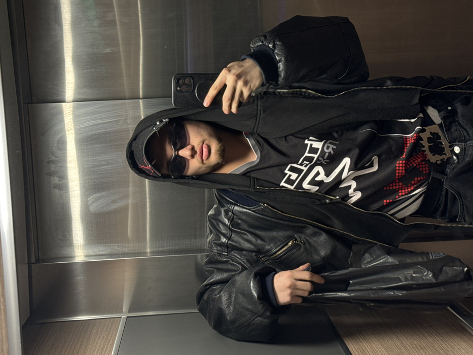 The image captures a single male individual taking a selfie inside an elevator in Asnières-sur-Seine, France. The subject, appearing to be in his late teens or early twenties, has short facial hair on his chin and wears dark, oval-shaped sunglasses. He is dressed in a black backward baseball cap with red detailing and metal studs, a black hooded sweatshirt with the hood up, a black leather bomber jacket, and a black t-shirt with a white and red graphic design. He also wears dark trousers with a prominent metallic belt buckle featuring a jagged outline and intricate design. In his right hand, he holds a black smartphone, positioned to capture the image. His left hand holds a dark, crumpled plastic bag, resting against his left side. The elevator interior consists of brushed metallic panels, exhibiting vertical streaks and reflections, with a narrow vertical panel of light wood-grain material visible on the left.