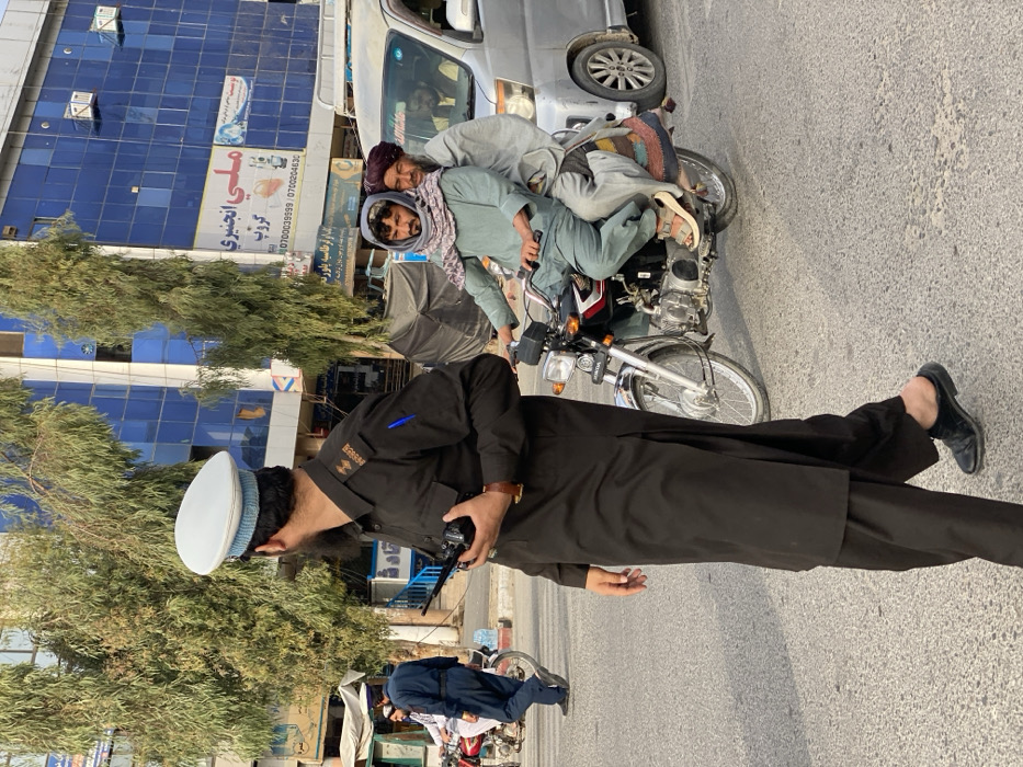 The image portrays an urban street scene in Kandahar, Afghanistan, under clear daylight conditions. In the foreground, a man, identified by his uniform and shoulder patch reading "KANDAHAR TRAFFIC POLICE," walks towards the right, looking over his right shoulder. He wears a dark brown tunic and wide-legged pants, a light blue and white peaked cap, black slip-on shoes, and a brown wristwatch on his left wrist. He holds a black handheld radio in his right hand, and a blue pen is visible in his left breast pocket.
To the right of the foreground officer, two men are riding a red and chrome Honda motorcycle. The driver, a younger man with a beard, wears a light green tunic, light-colored pants, a grey and white patterned head covering, and sandals. The passenger, an older man, sits behind him on a patterned textile draped over the seat, wearing a light grey tunic and a maroon turban-like head covering.
In the background, a multi-story building with blue reflective glass windows is prominent. A large sign on this building displays "ملي انجینری گروپ" (Milli Engineering Group) in Dari/Pashto, along with Afghan phone numbers (0700039999, 0700204630). Tall, slender trees with light green foliage are situated between the street and the buildings. Further left in the background, another individual in a dark uniform walks beside a red motorcycle. To the far right, a silver minivan or small bus is partially visible, with at least two individuals inside. The street surface is paved asphalt.