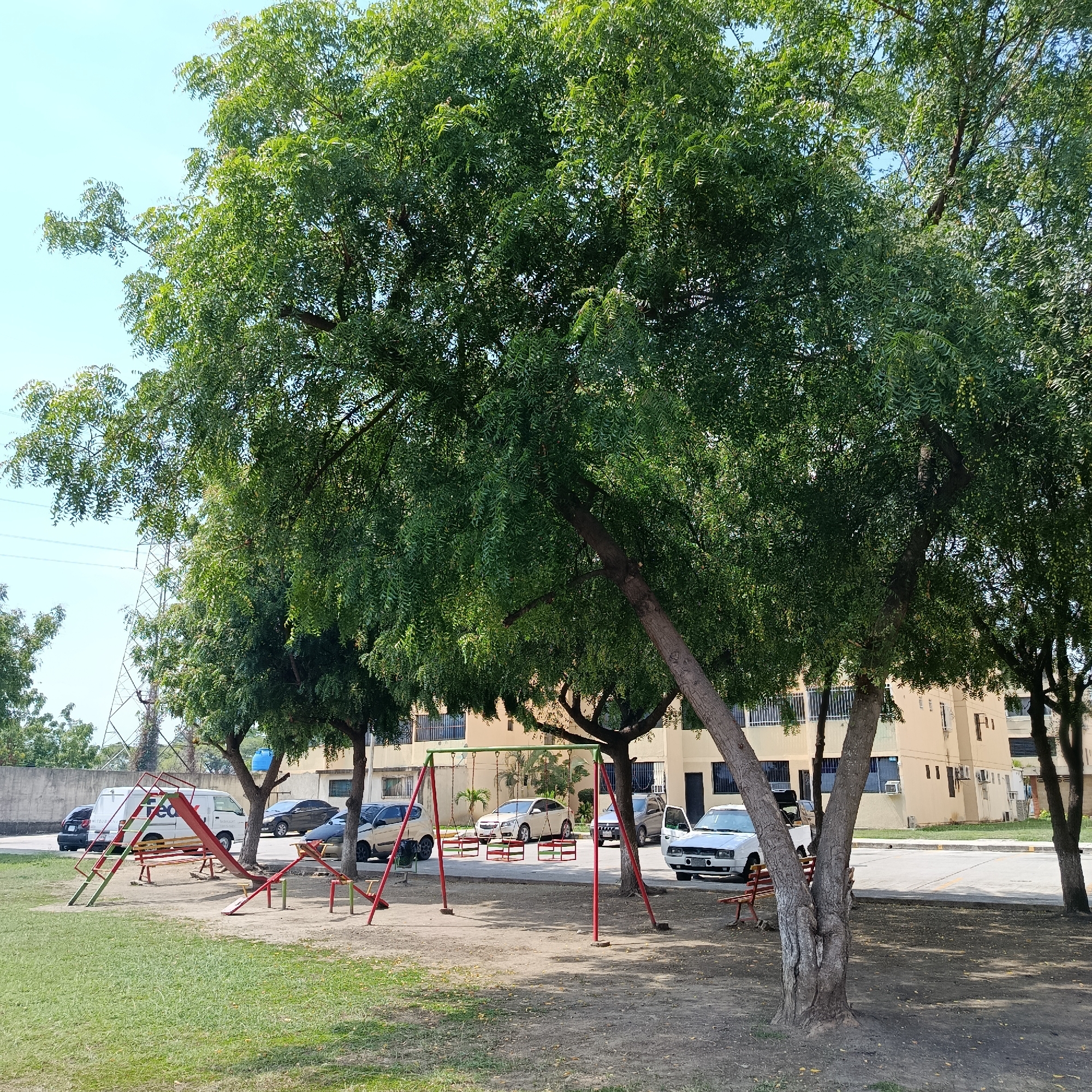 The image shows a park in Los Guayos, Venezuela. The park contains playground equipment, including a slide, a seesaw, and swings. Several trees provide shade over the park. Behind the park, there is a building and a parking lot with several parked cars. In the background, power lines are visible. The sky is clear and blue.
