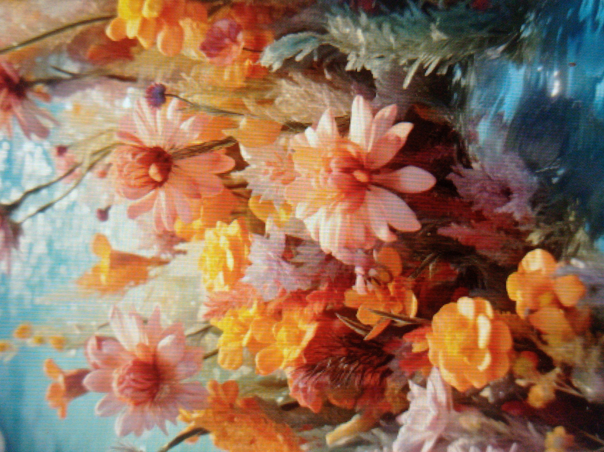 The image displays a close-up, slightly blurred view of a vibrant bouquet of flowers. The flowers are predominantly in shades of orange, pink, and yellow, with some lighter pink and red accents. Several types of flowers are visible, including those with layered petals that resemble chrysanthemums or dahlias, and others with more delicate, elongated petals. Green foliage is interspersed throughout the bouquet. The background is a bright, textured blue, suggesting an out-of-focus sky or water. The lighting appears bright, casting soft shadows and highlighting the textures of the petals. There are no people or discernible urban elements in the image, making it impossible to determine any connection to Mukachevo, Ukraine, or any specific city. The scene is solely focused on the arrangement of flowers.