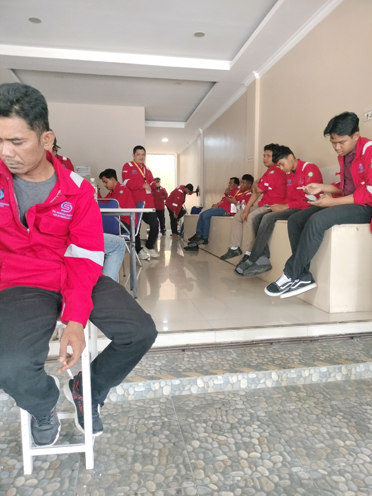 In Batam, Indonesia, a group of men in red uniforms are seated and standing in an indoor waiting area. The individual in the foreground, seated on a white stool, is wearing a red jacket with a company logo and black pants, and appears to be holding a cigarette. Behind him, several other men in similar red uniforms are seated on a long bench, some looking at their phones or talking. The environment includes beige walls, white trim, and a tiled floor with a cobblestone pattern on the steps leading out.