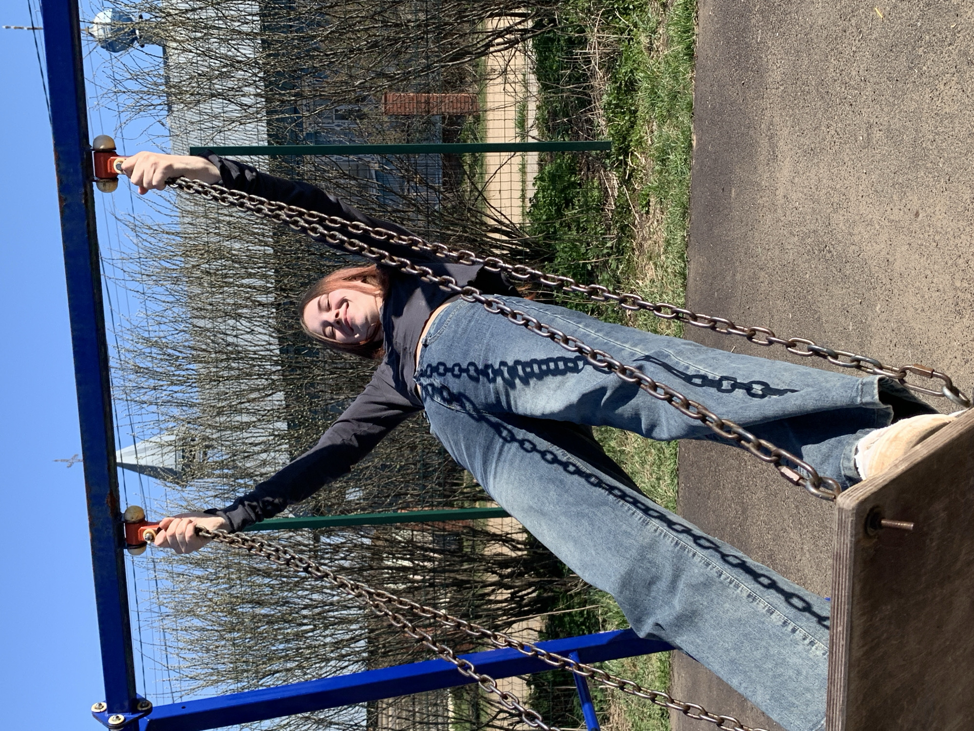 A young woman in blue jeans and a dark long-sleeved shirt smiles while holding the chains of a swing set. The swing set has a blue frame and a wooden seat. The background includes trees with bare branches, a metal fence, grass, and a paved path. The location appears to be a playground or park. The city is not evident from the image.