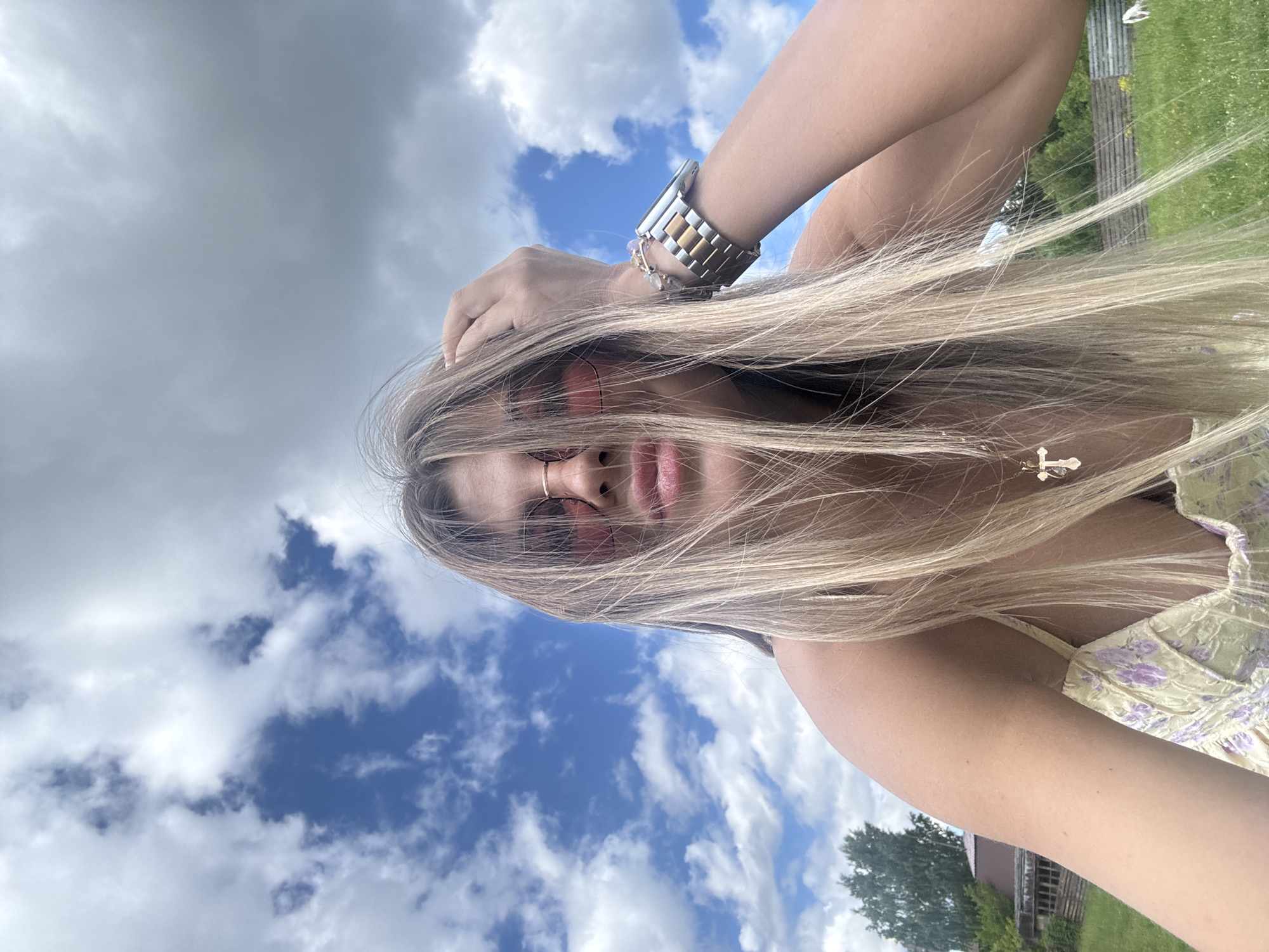 The image is a selfie taken outdoors, likely in a rural area, with a woman in the foreground. She has long blonde hair and is wearing pink-tinted sunglasses and a light-colored dress with a floral pattern. Her left hand is raised and touching her head. She is wearing a watch on her left wrist. In the background, there is a cloudy blue sky, green grass, trees, and a wooden structure. The location is specified as Пастухово, Russia.