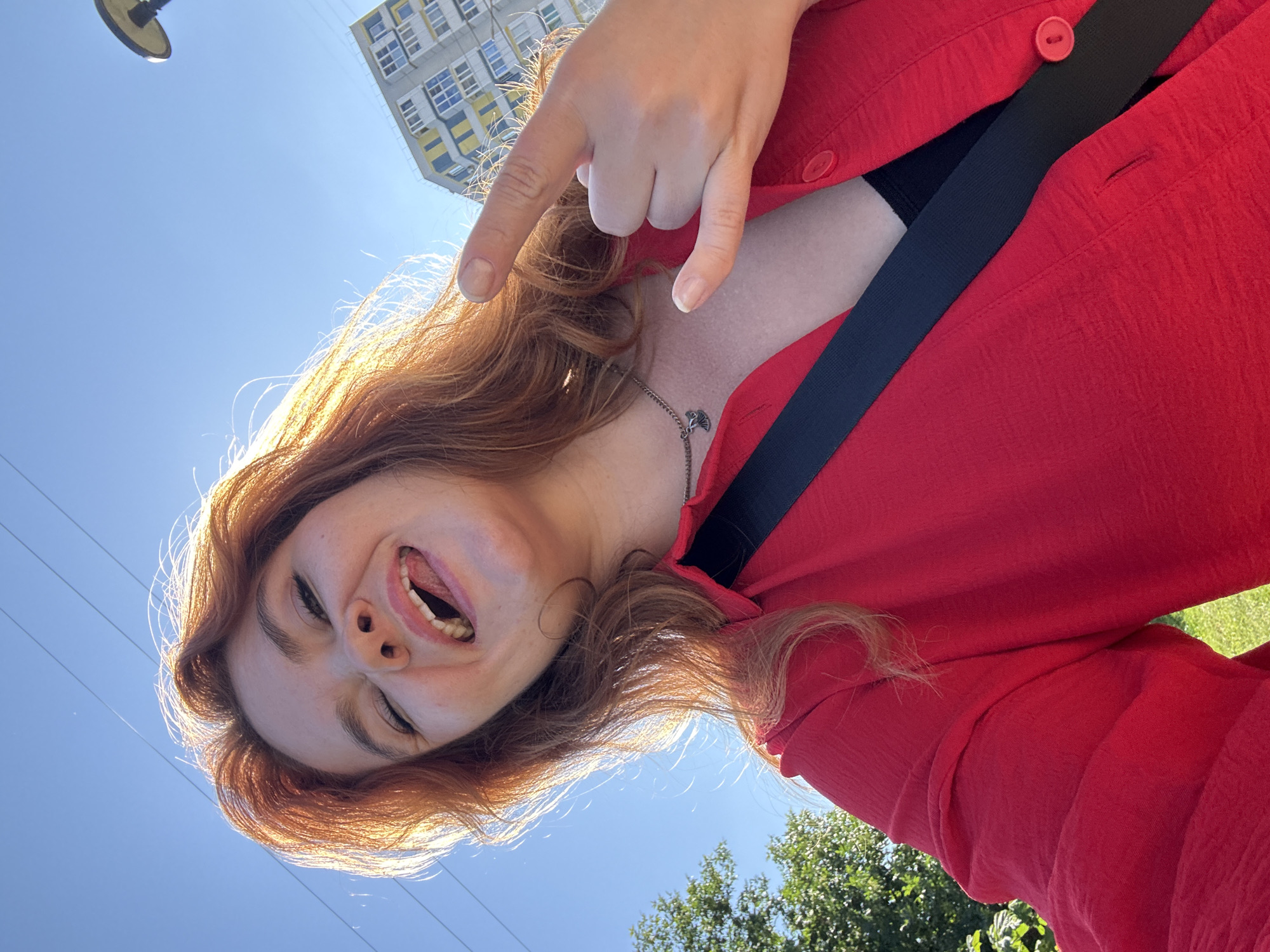 A young woman with auburn hair is taking a selfie outdoors on a sunny day. She is wearing a red collared shirt with a black strap across her chest and a black top underneath. Her tongue is sticking out and her eyes are closed in a playful expression. Her right hand is raised, forming a rock and roll hand gesture. In the background, there is a blue sky, a modern apartment building with yellow and blue accents, green trees, and a lamppost. The lighting suggests it is daytime, and the presence of a building and lamppost indicates an urban or suburban setting.