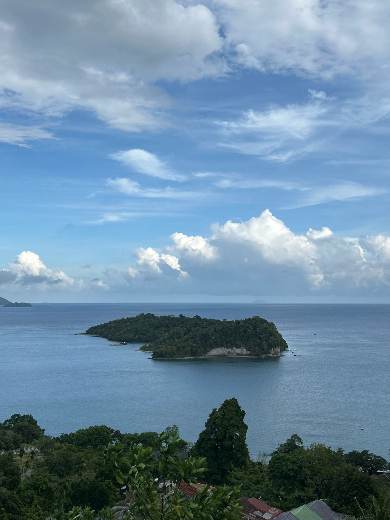 A panoramic view captures a lush, green island situated in the calm, blue waters of the ocean. The island is densely covered with trees and features a rocky cliff face. The sky above is a vibrant blue, dotted with fluffy white clouds. In the foreground, more trees and the rooftops of houses are visible, suggesting a coastal settlement on the outskirts of the city of Sabang, Indonesia. The overall scene evokes a sense of tranquility and natural beauty.