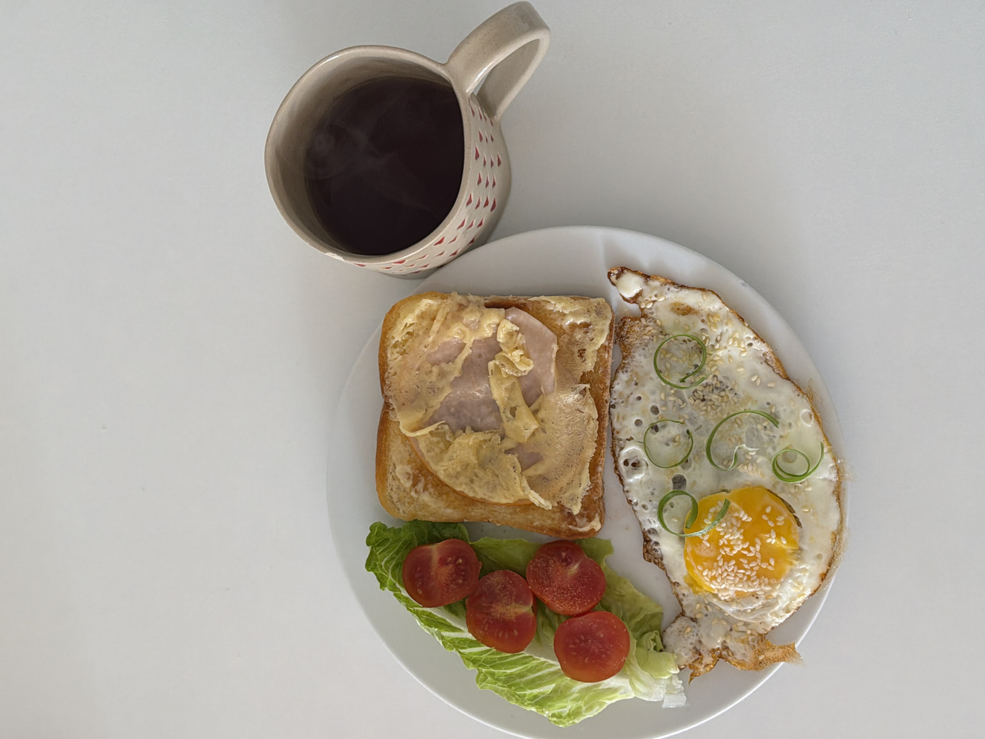 An overhead shot displays a breakfast or brunch meal set on a plain white surface, likely a table or countertop. The primary subjects are a white ceramic mug and a white round plate filled with food.
The mug, positioned in the upper left, is light-colored and features small, irregular red markings, possibly hearts or dots, on its exterior. It holds a dark beverage, from which faint steam is visibly rising, suggesting it's hot coffee or tea.
On the plate, arranged neatly, are several food items. To the left, a slice of golden-brown toasted bread is topped with a slice of ham and a layer of melted cheese or butter. Below the toast, a vibrant green lettuce leaf forms a bed for four halved cherry tomatoes. To the right of the plate, a fried egg is the star, featuring a bright yellow, runny yolk and slightly crispy, browned edges. The egg is garnished with a sprinkling of white sesame seeds and delicate curls of thinly sliced green onion.
The overall presentation is simple and clean, suggesting a homemade meal. The bright, even lighting implies an indoor setting, possibly during the morning or early afternoon. There is no visible text in the image. Although not discernible from the image itself, this meal is situated in Tel-Aviv, Israel, according to the provided context.