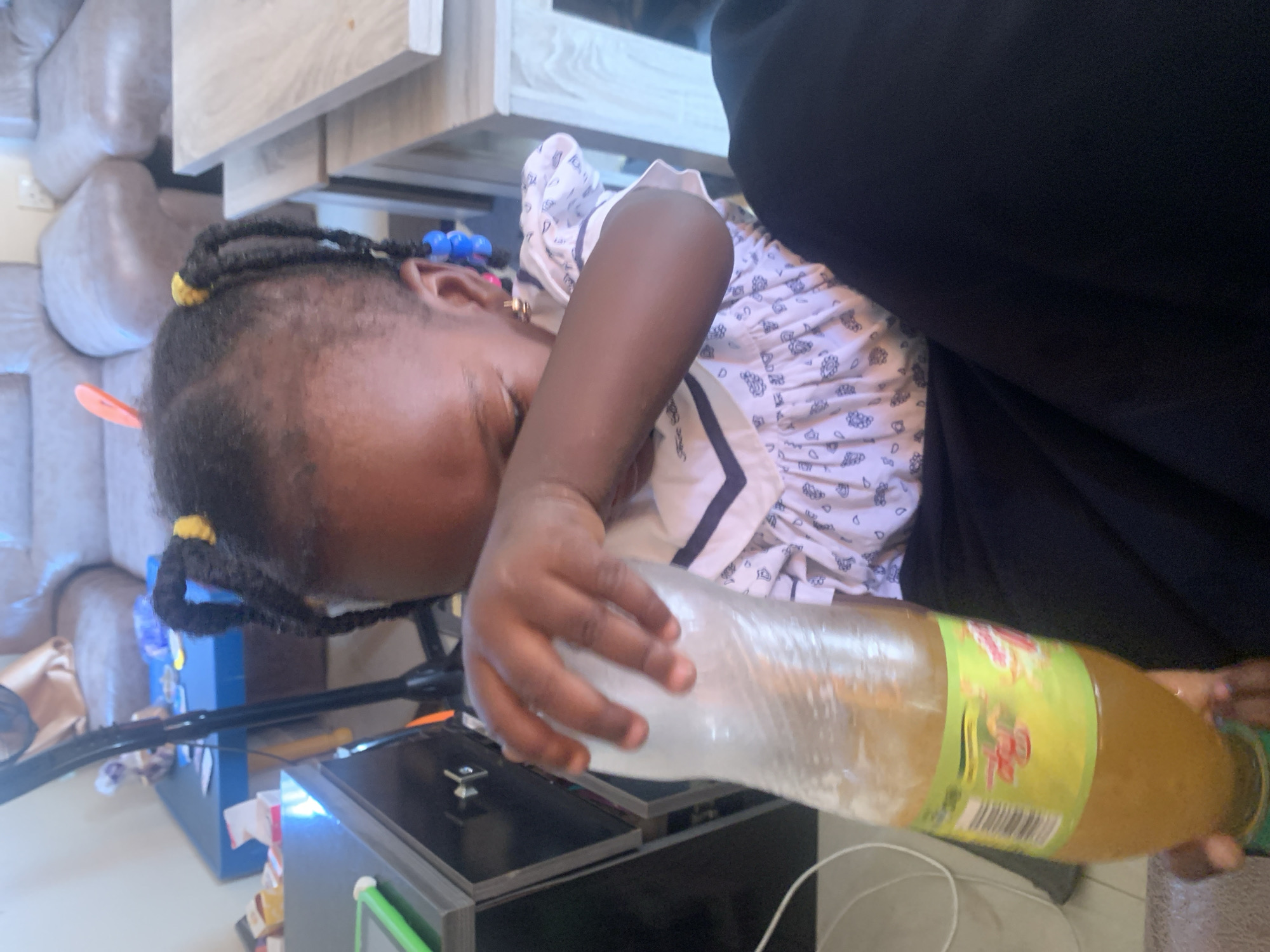 In a dimly lit indoor setting, a young Black girl is the primary subject. She is lying down, facing to her right, with her left arm extended. Her hair is styled in braids with yellow and blue beads. She is wearing a white dress with a pattern of blue outlines. She is holding a transparent plastic bottle filled with an amber liquid, possibly a beverage. Her hand is gripping the bottle.
The background shows parts of a living room. There are upholstered sofas in a neutral tone, and a wooden shelf or cabinet is visible. On the floor, there is a black electronic device with some cables and a green accent. Another blue object with stickers is also partially visible. The overall impression is casual and domestic.
The image seems to have been taken during the day, but the indoor lighting creates a subdued atmosphere. There are no clear indicators of the weather or specific time of day beyond it being indoors. The girl's expression is difficult to discern due to her orientation and the lighting, but her focus appears to be on the bottle she is holding.
A label on the bottle is visible, featuring green and yellow colors and some text in what appears to be Latin script, though it is not clearly legible. The setting suggests a home environment in Gura Top, Nigeria.