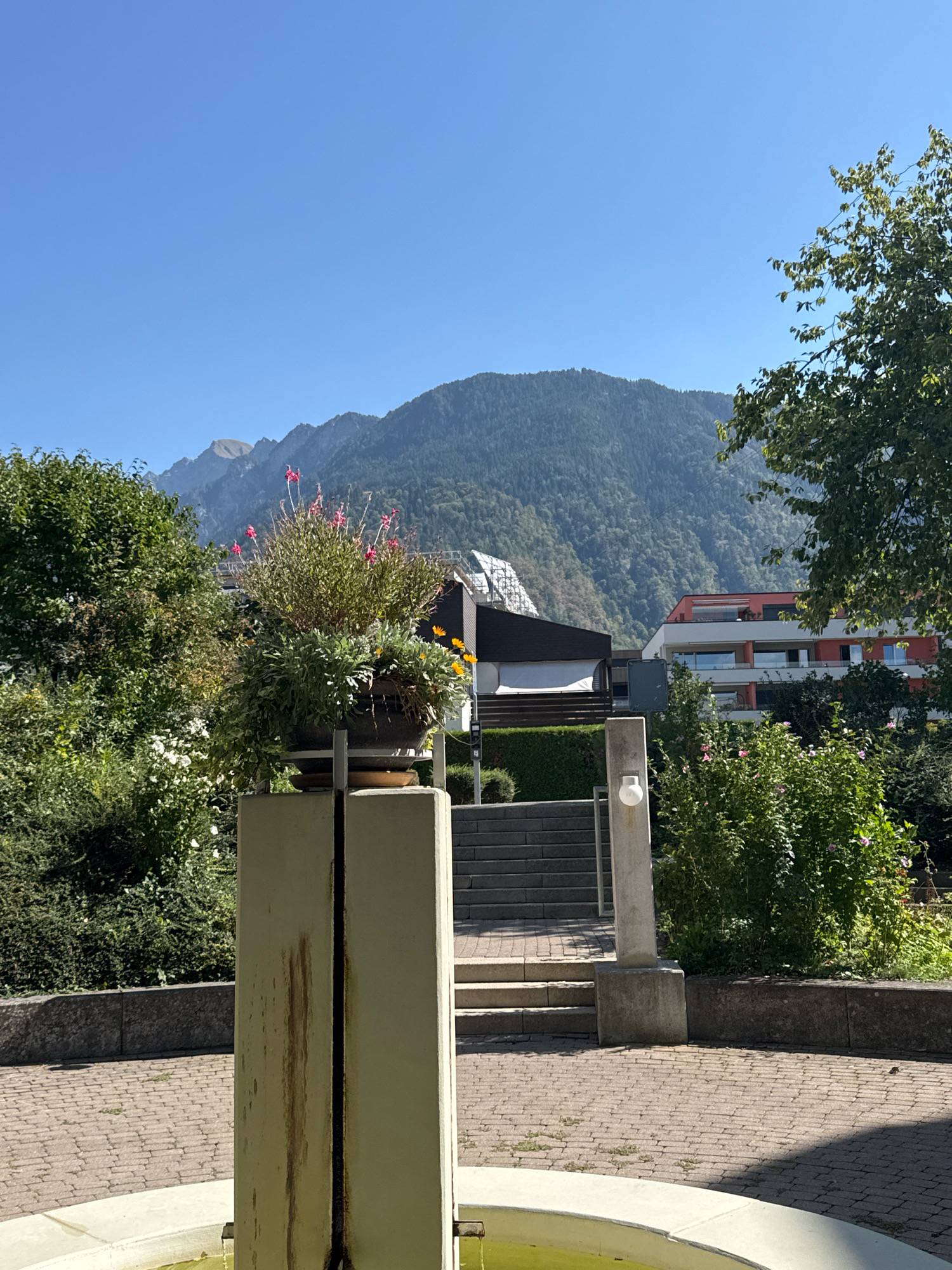 A wide-angle, eye-level shot shows a scenic view of a town or city nestled at the base of a lush, green mountain range under a clear blue sky. In the foreground, a round, white fountain basin is partially visible, with a light green liquid inside. A light-colored, rectangular pillar stands in front of the fountain, displaying a dark, vertical stain down its center. A large potted plant filled with vibrant pink and yellow flowers sits atop a similar pillar to the right of the first.
The middle ground features a paved walkway leading up a set of concrete stairs to a building with a modern, red and white facade. Behind this building, a dark, angled roof structure is visible. Green trees and bushes frame the scene, with a large tree on the right side of the frame casting a shadow on the walkway. The background is dominated by the imposing, verdant mountain.