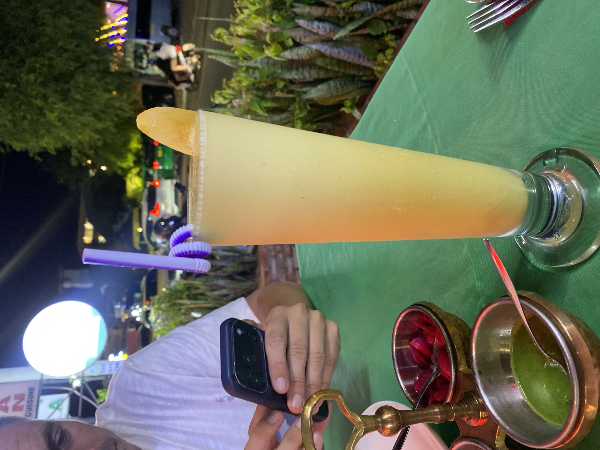 In Phu Thuy Ward, Vietnam, a person in a white t-shirt is sitting at a table covered with a green tablecloth, holding a smartphone. In front of them is a tall glass of a yellow, frothy beverage, possibly a mango lassi, garnished with a slice of mango and a purple swirled straw. Two small brass bowls containing red pickled items and a green sauce, each with a spoon, are also on the table. In the background, illuminated by streetlights, cars are visible, along with lush green foliage and building lights, indicating an outdoor setting at night.