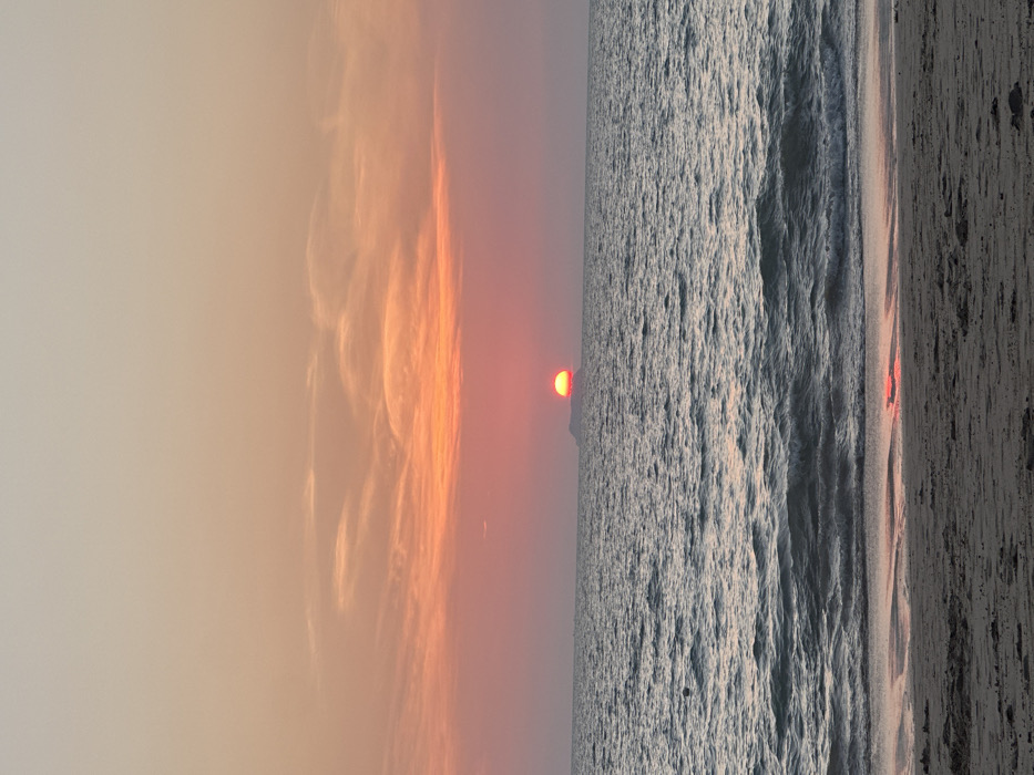 The image depicts a sunset over a body of water at Phra Ae beach in Phra Ae, Thailand. The sky exhibits a gradient of colors, transitioning from light peach and pale orange at the top to a vibrant orange and red hue near the horizon. Elongated, wispy clouds are present, primarily in the middle section of the sky.
The sun, a distinct orange-red circular orb, is partially visible and appears to be setting behind a low, dark landmass on the distant horizon. This landmass forms a silhouette against the bright sun.
Below the horizon, the water's surface is a dark blue-grey, characterized by ripples and small breaking waves. A faint, reddish reflection of the sun is visible on the water closer to the foreground. A small, dark, circular object, possibly a buoy, is discernible floating on the water in the mid-ground to the left of the frame.
The foreground consists of a sandy beach, appearing dark brown-grey. The sand shows variations in texture and color, indicating both dry and wet areas, with the wet sand closer to the water's edge. Minor indentations or marks are visible on the sand. No people, major objects, or significant actions are depicted in the scene.