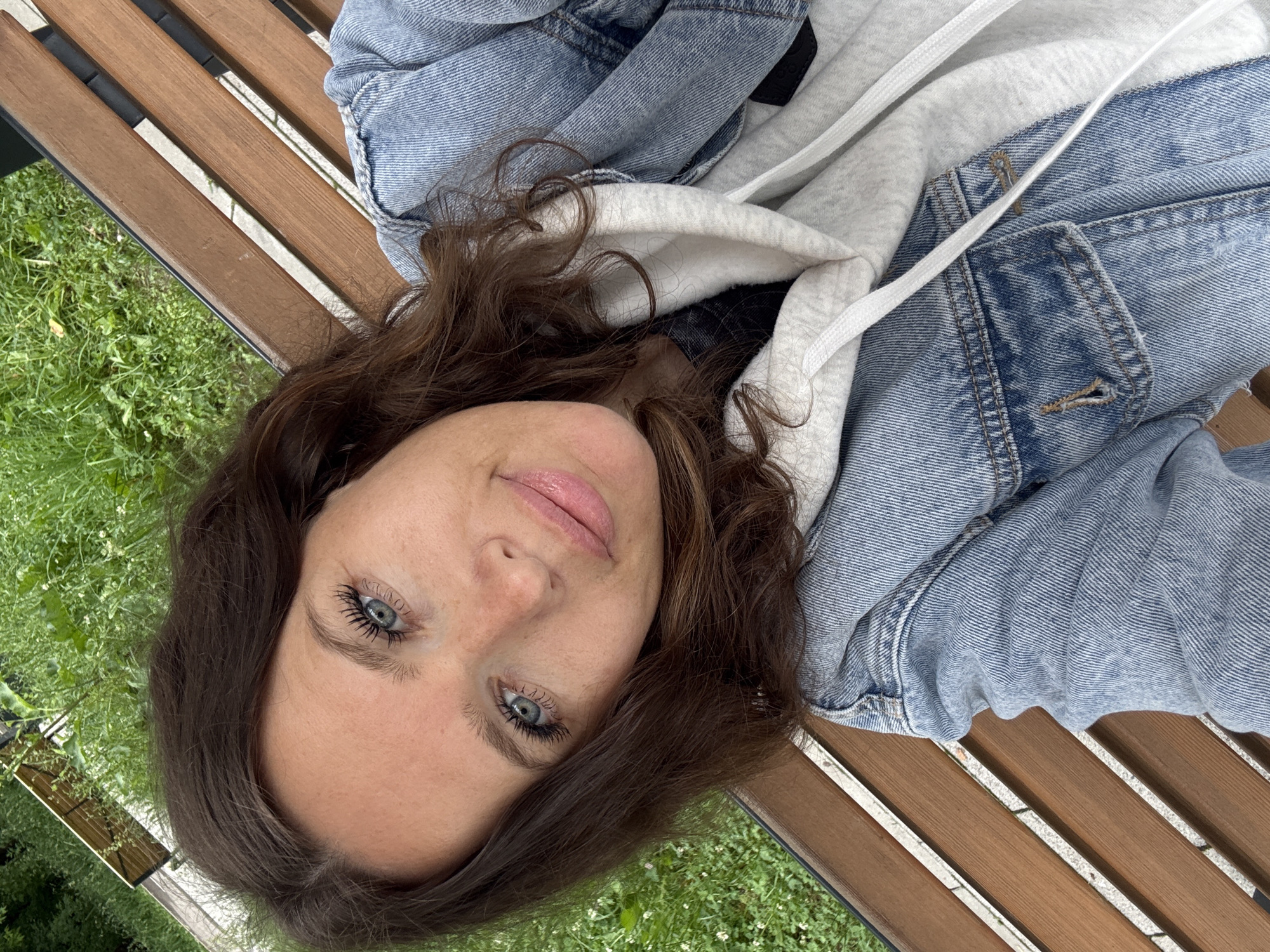 A woman is taking a selfie while sitting on a wooden park bench. She has brown hair, blue eyes, and is wearing a denim jacket over a gray hoodie. The background shows green grass and foliage, suggesting an outdoor park setting. There is no visible indication of the location being in Moscow, Russia.