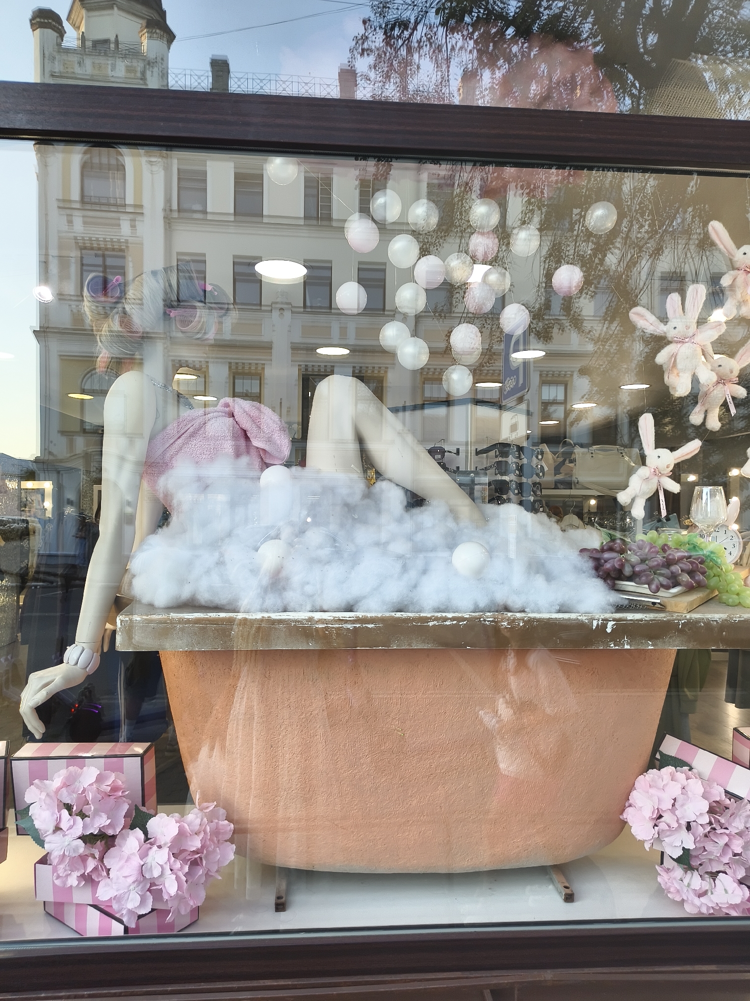 This image shows a store window display in an urban environment. The display features a mannequin with hair rollers and a pink top, positioned as if lounging in a bathtub filled with white fluffy material resembling clouds or bubbles. The bathtub is a coral-orange color. There are also decorative pink hydrangeas in striped boxes and plush white bunnies hanging from the ceiling. The background of the window shows a reflection of buildings and trees, suggesting the display is on a street in a city. The lighting in the scene is mixed, with artificial lights from within the store and natural light from outside, creating reflections on the glass.