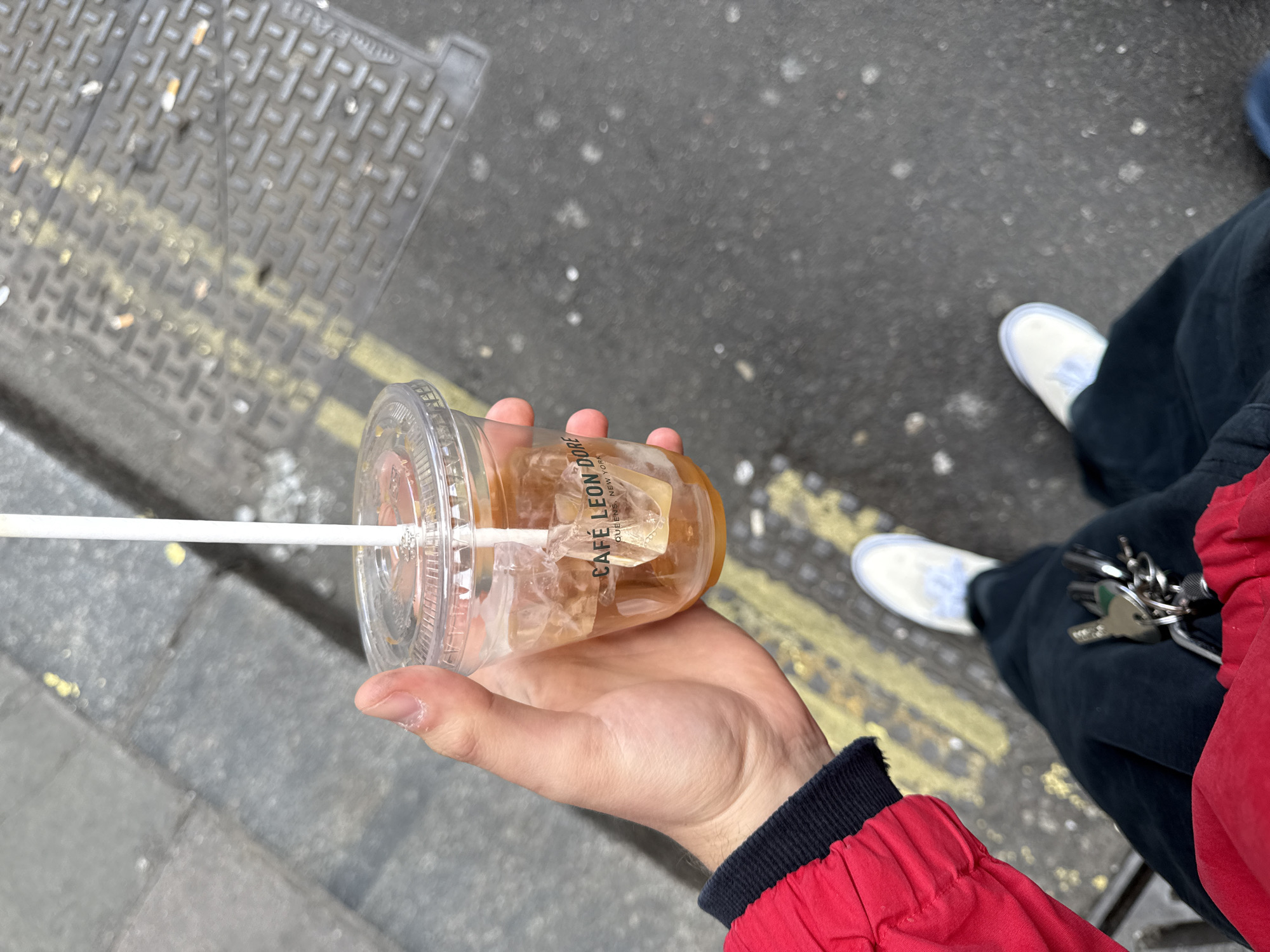 The image shows a person holding a clear plastic cup of iced coffee with a white straw, with the logo "CAFE LEON DORE, QUEENS, NEW YORK" printed on the cup. The person is wearing a red jacket with a black trim. The background includes a street with a metal grid pattern, yellow painted lines, and asphalt. In the lower right, the person is wearing black pants and white sneakers. There is a set of keys attached to their pants. Given the cafe branding, which is New York-based, the location is unlikely to be London, United Kingdom, but rather somewhere in the USA.