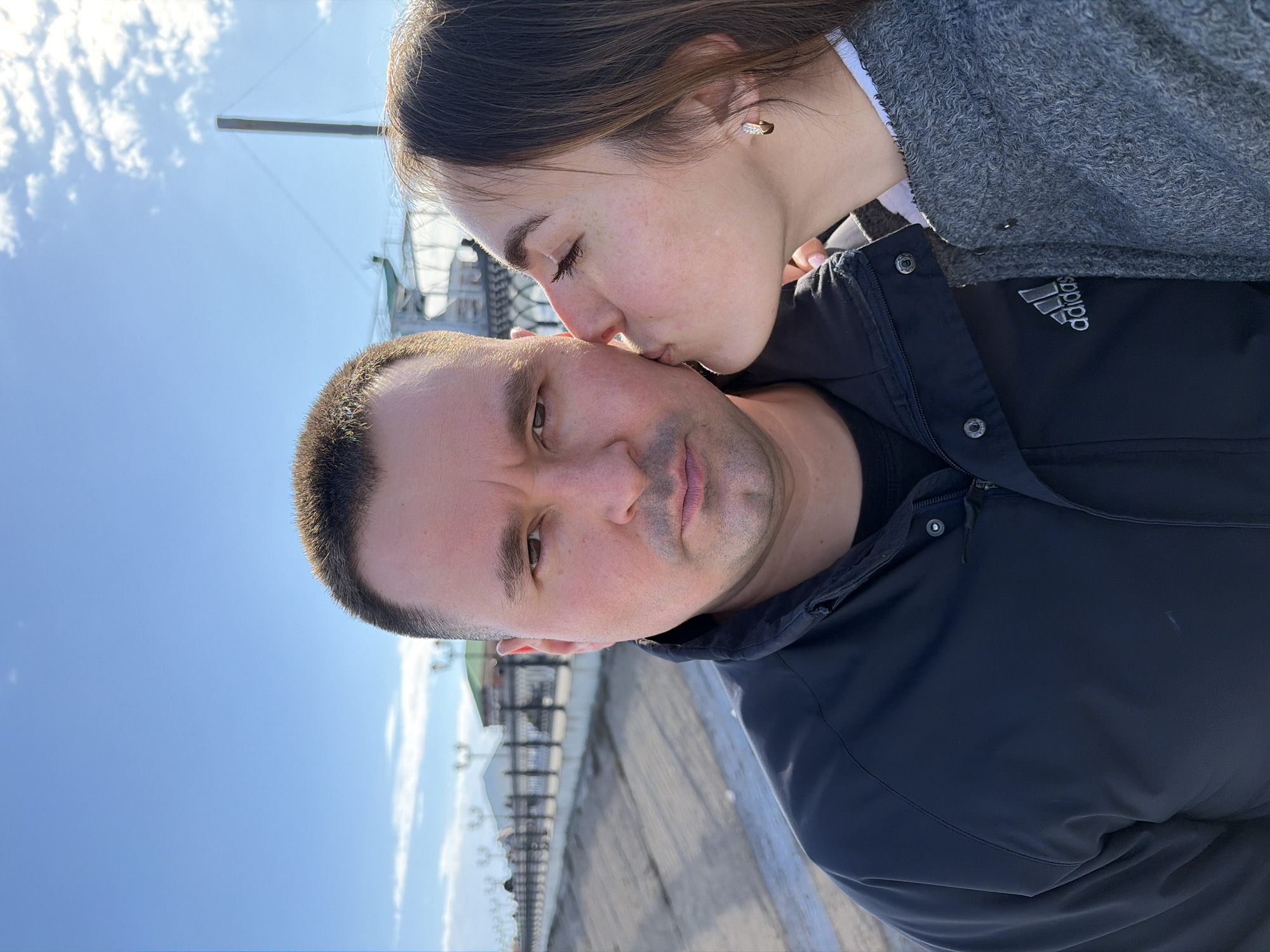 A man is being kissed on the forehead by a young woman in an outdoor setting. The woman is wearing a gray sweater and has an earring visible in her left ear. The man is wearing a black jacket. They are standing on what appears to be a wooden pier or walkway, with a view of a fence or railing in the background and a bright sky above. Based on the visible architectural style in the background, this is possibly the location of the pier in Sarapul, Russia.