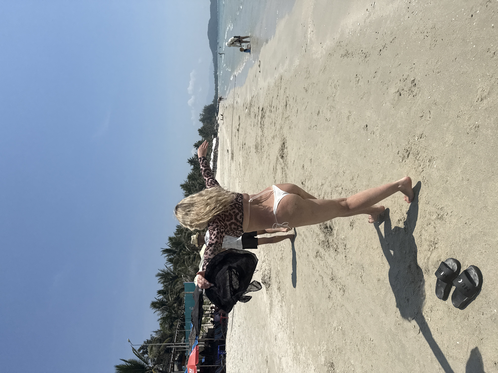 On a bright, clear day in Dong Ninh Hoa Ward, Vietnam, a woman stands on a sandy beach, facing away from the camera. She is wearing a swimsuit bottom and a long-sleeved top with an animal print. She has long, blonde hair and her arms are outstretched. In her hands, she holds a large black hat. Behind her, a person is walking on the beach. The shoreline, water, and some other beachgoers are visible in the distance. Palm trees and some structures are seen to the left, along with other beachgoers. A pair of black sandals sits on the sand in the foreground.