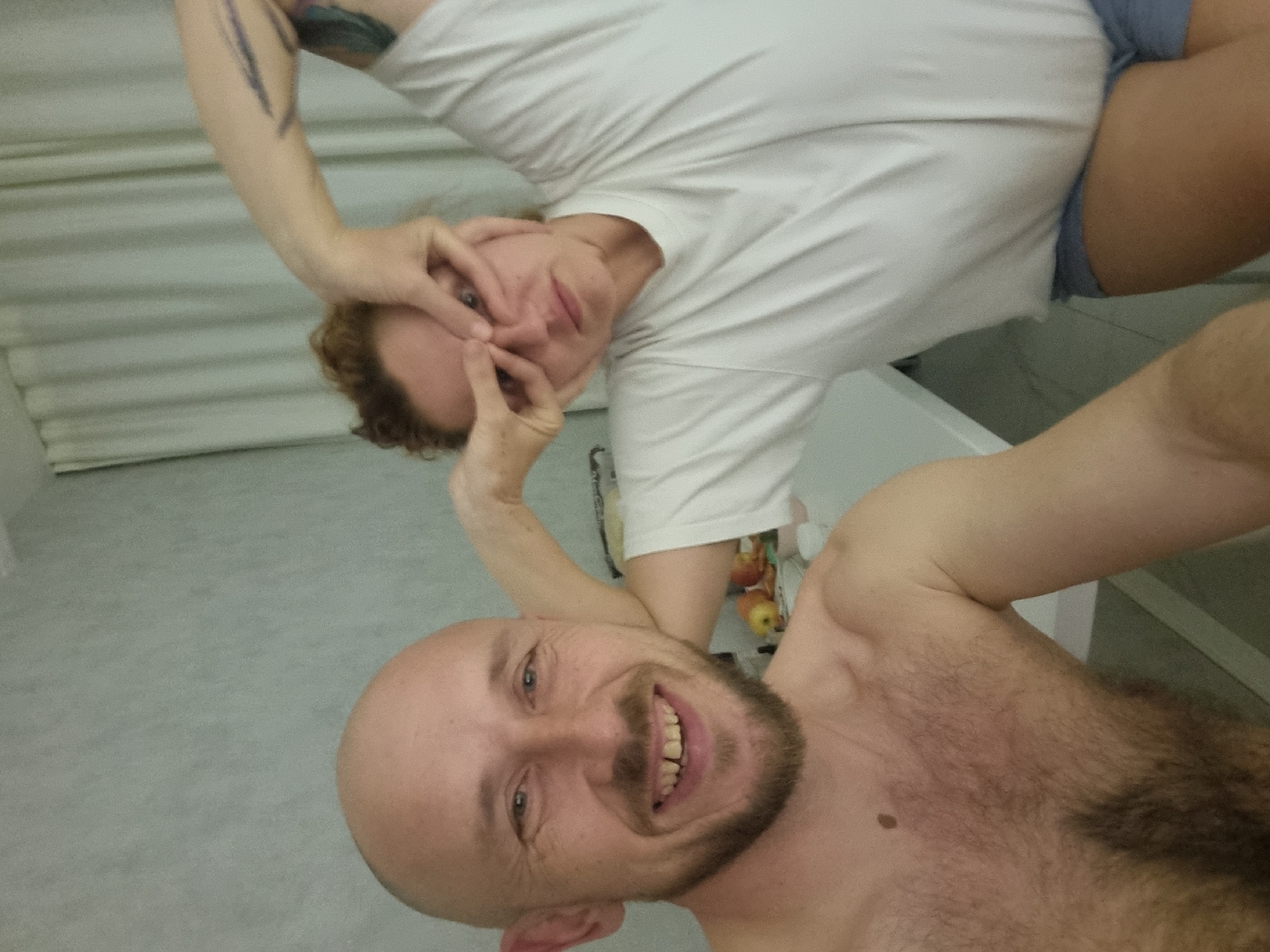 A selfie depicts two people in an indoor setting. The foreground features a bald man with a beard, smiling at the camera. He is shirtless, with chest hair visible. Above him, a woman with curly reddish-brown hair wears a white t-shirt and blue shorts. She has her hands near her eyes, making a face. The background appears to be a simple interior space, possibly a room in a house or apartment. The precise location within the city of Sochi, Russia, is not identifiable from the image.