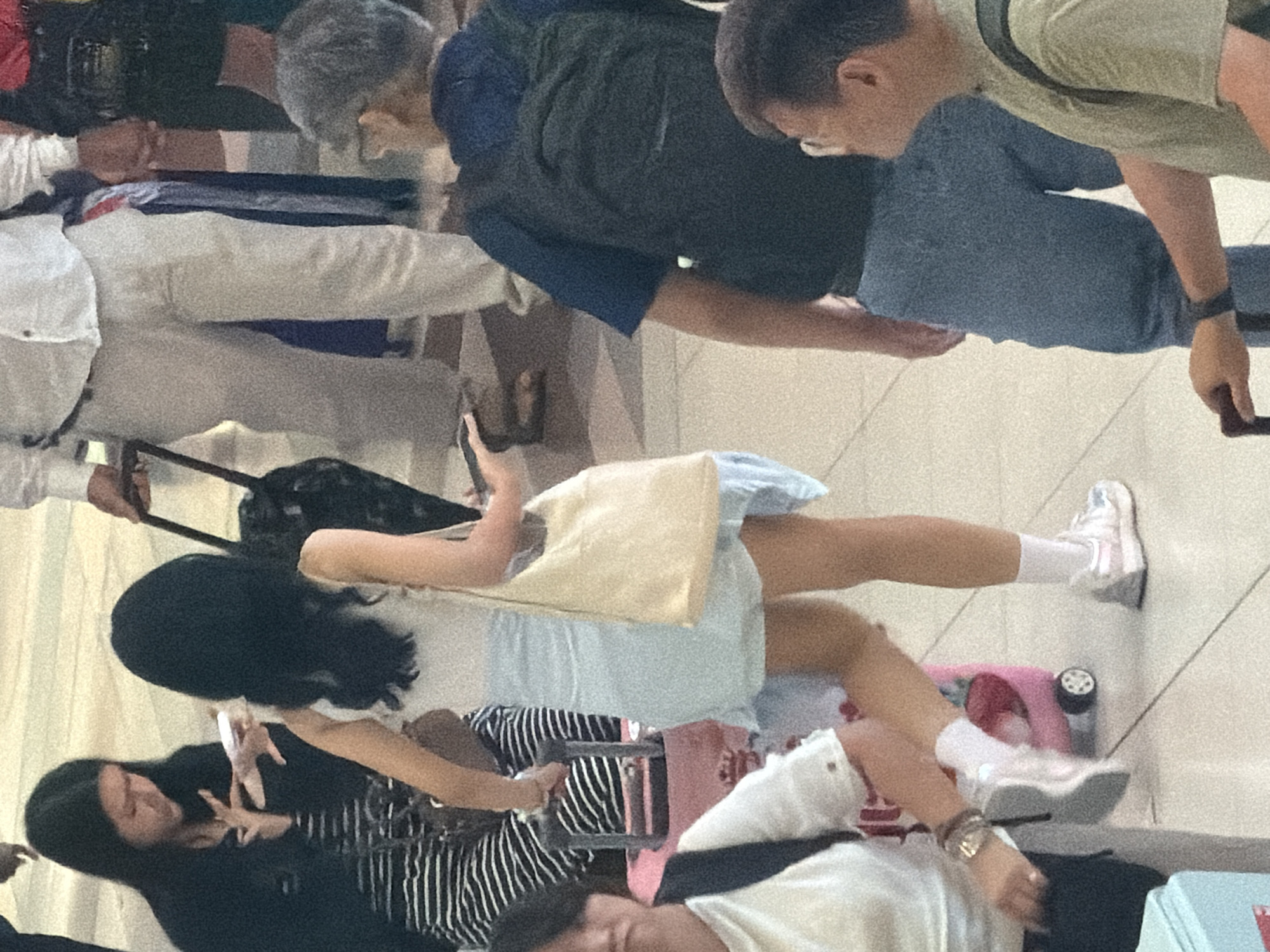 In Nong Prue Subdistrict, Thailand, a group of individuals are present in a public indoor area, possibly an airport or large transit hub, characterized by a tiled floor and muted lighting. A woman in the foreground is walking, wearing a light blue dress and carrying a cream-colored shoulder bag. She has long dark hair. To her left, another woman is seated, wearing a black jacket over a striped top, and is looking down at her hands. To the right of the walking woman, a man wearing glasses and a navy blue shirt with a backpack is walking away from the camera, and a man in a light green shirt is holding a piece of luggage. Further in the background, individuals in white shirts and light-colored pants are visible, as well as luggage. The scene depicts everyday movement and travel within a busy public space.