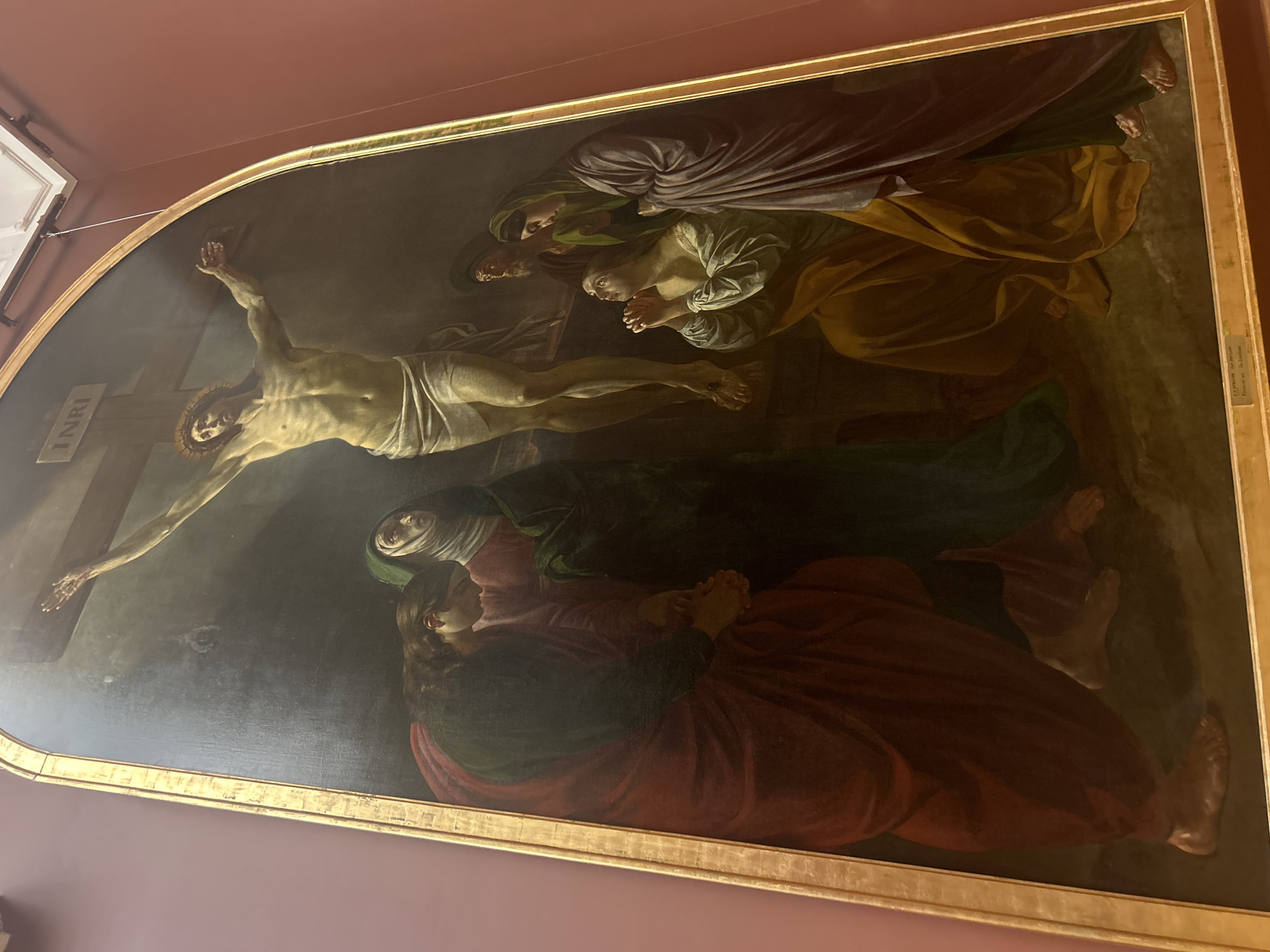 This is a painting of the crucifixion of Jesus Christ, displayed in the State Hermitage Museum in Saint Petersburg, Russia. The painting is housed in a large, ornate gold frame, and is set against a dark, somber background. Jesus is depicted on the cross, with a loincloth wrapped around his waist. His body is emaciated and bruised, with a crown of thorns on his head.
Below the cross, several figures are gathered, representing mourners. On the left, a woman in a deep red robe and a green head covering is depicted with her head bowed and hands clasped. To her right, another woman in a dark green robe and a white head covering gazes upwards with a mournful expression. Further to the right, a woman in a light blue and yellow robe is kneeling with her hands clasped in prayer, looking up towards Jesus. Behind her, a man with a green turban and a woman in a purple robe stand with somber expressions. The overall mood of the painting is one of deep sorrow and religious reverence.