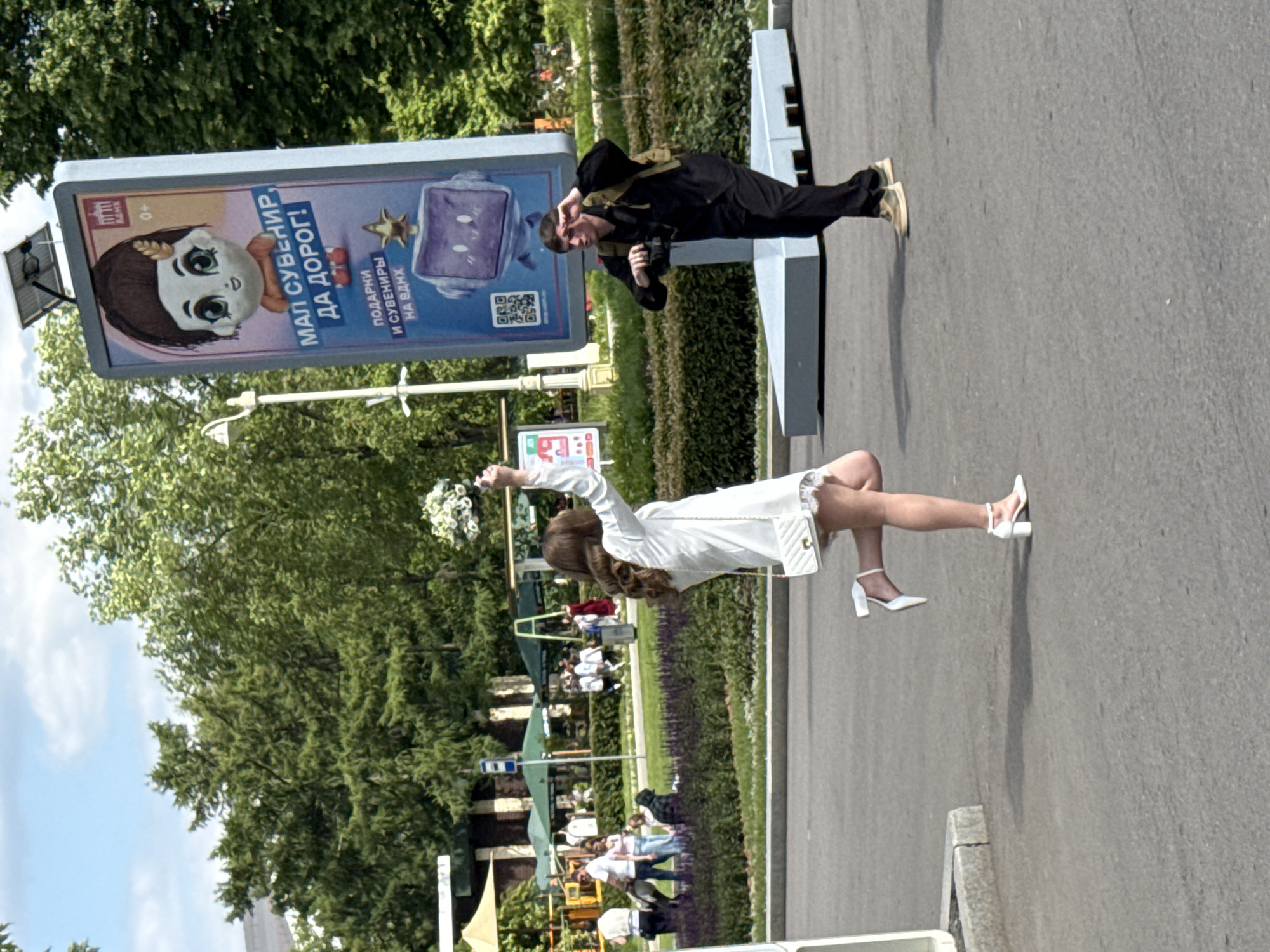 The image depicts an outdoor scene, likely a park or public walking area, under bright daylight. In the foreground, on the right, a young woman in a white long-sleeved top, short white skirt, and white block-heeled shoes is captured in a dynamic pose, her left arm raised as she holds a white floral bouquet. She carries a small white quilted handbag.
To her left, in the middle ground, a man dressed in black, possibly a jumpsuit, with beige boots and a beige harness, stands next to a large advertising display. He holds a camera at his side, looking towards the woman, suggesting he is photographing or filming her. He also wears a backpack. Behind him is a modern, light-grey angular bench or structure.
The prominent advertising display features a cartoon illustration of a young girl with large eyes and braided hair, holding what appears to be a hot dog. Cyrillic text on the billboard reads: "МАЛ СУВЕНИР, ДА ДОРОГ!" (A small souvenir, but precious/expensive!), and below it, "ПОДАРКИ И СУВЕНИРЫ НА ВДНХ" (Gifts and souvenirs at VDNKh). An age restriction "0+" and a QR code are also visible.
The background shows lush green trees and foliage, with several indistinct figures of people further in the distance, some under shaded structures, indicating a busy public space. The clear sky and strong shadows suggest the time of day is midday or early afternoon. The scene conveys a celebratory photoshoot taking place, likely at VDNKh in Moscow, Russia, as indicated by the billboard text.