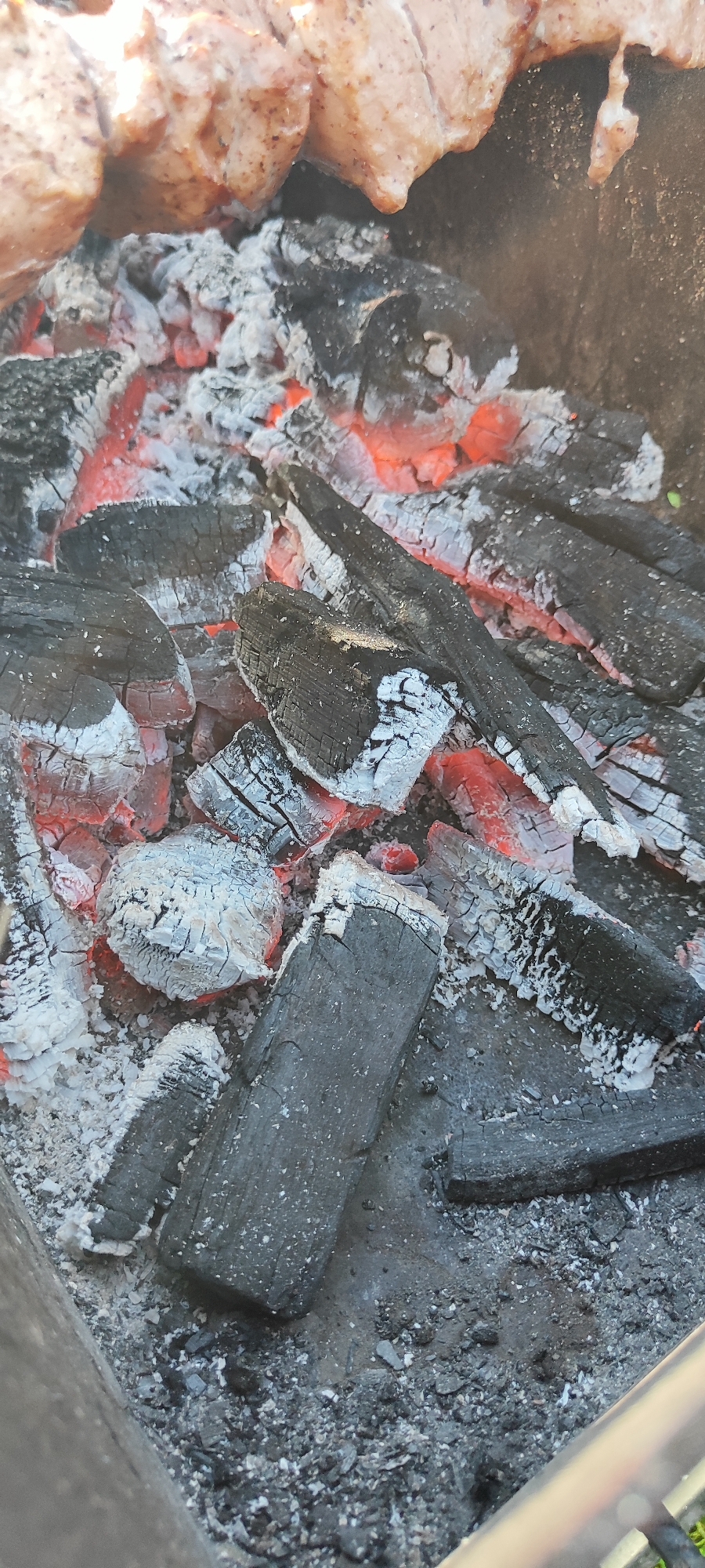 This image shows a close-up view of hot coals and ashes in a grill. The coals are glowing red in several areas, indicating high heat. The charred wood pieces are covered in a layer of white ash, typical of burnt material. In the upper portion of the image, partially visible, are pieces of raw meat skewered, suggesting that food is being prepared for grilling. The scene appears to be outdoors, possibly in a backyard or a designated grilling area. The image does not provide any specific information about the city of Лёзненскі сельскі Савет, Belarus, other than its potential context. There are no people or specific interactions visible in this image.