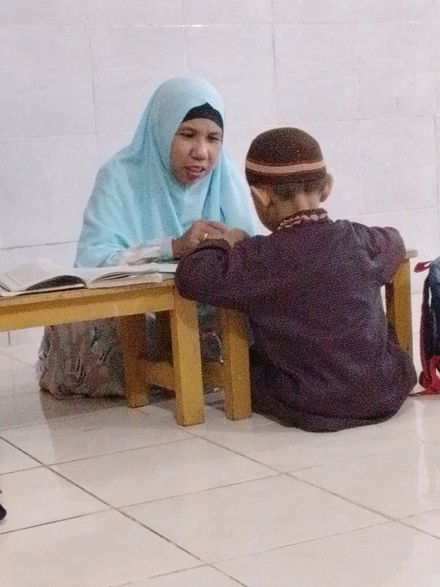 A medium shot shows an Indonesian woman wearing a light blue hijab and a young boy wearing a peci hat and a dark patterned shirt, sitting at a low, wooden table in a brightly lit room. The woman is on the left, facing the boy, with an open book in front of her. Her mouth is open as if speaking, and she is looking at the boy. The boy is on the right, facing away from the camera, with his back to the viewer. He is leaning forward, his hands on the table. The room has tiled floors and a white tiled wall in the background. A blue backpack with red straps is visible to the right of the table. The scene suggests an educational setting, possibly a lesson. The image quality is grainy.