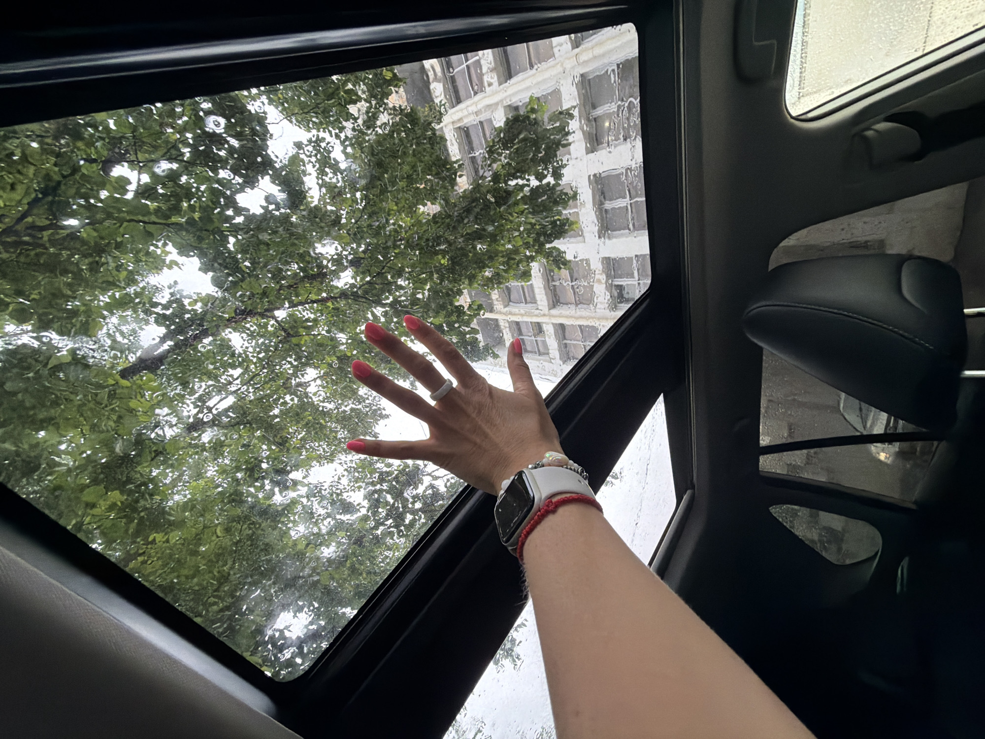 A person's left hand, adorned with bright pink nail polish and a ring, is raised towards the open sunroof of a vehicle. The sunroof reveals a view of lush green trees and a building with many windows, all seen through the rain-streaked glass. The person's wrist features a white smartwatch and a red beaded bracelet. The interior of the car, including a headrest and part of the dashboard, is visible in the foreground. The scene suggests a moment of connection with nature during inclement weather from within the car. The location is described as Birobidzhan, Russia.