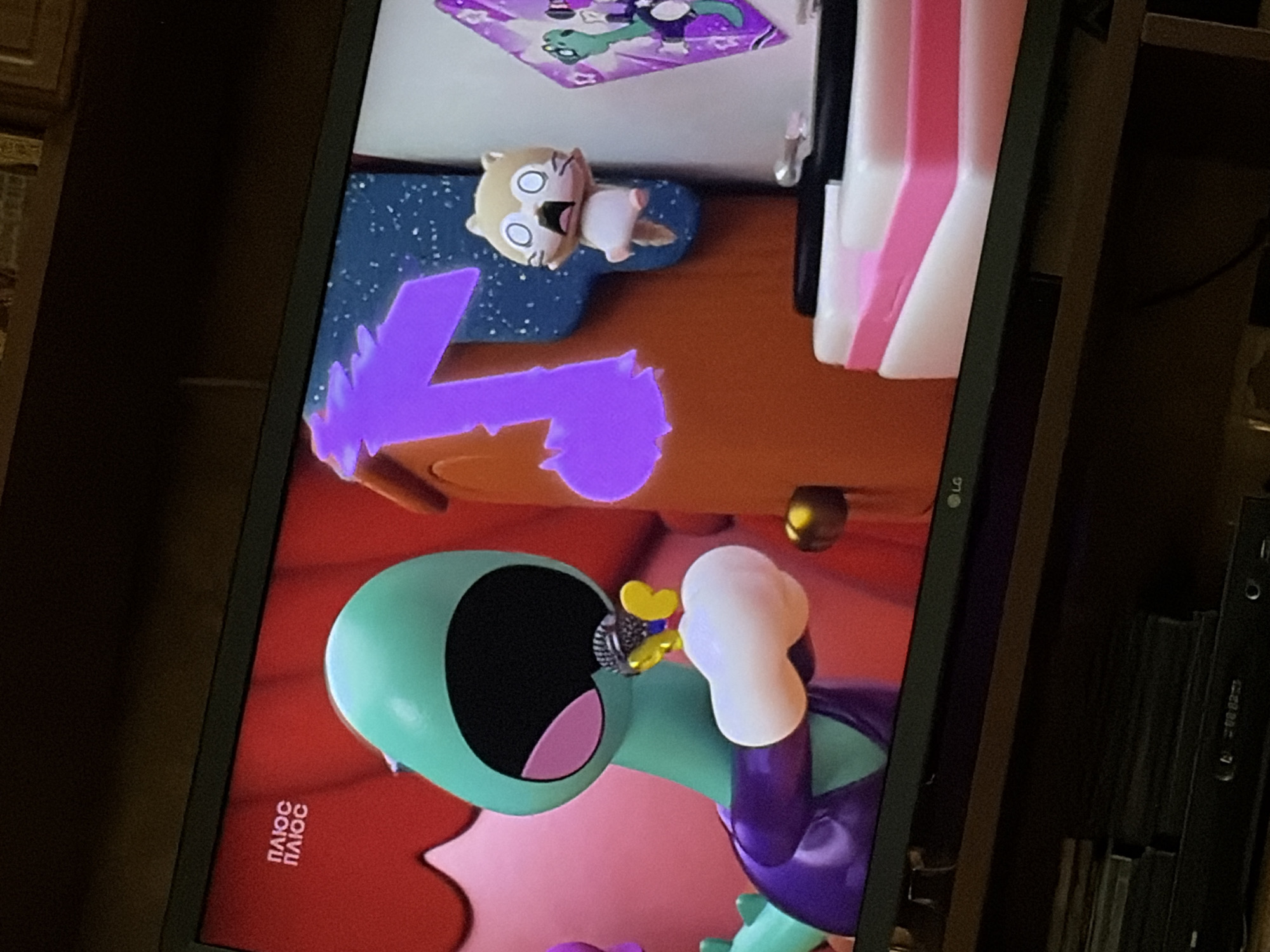 An eye-level medium shot captures a television screen displaying a colorful animated scene. In the foreground, a large, teal-colored, cartoonish creature with a wide-open mouth and pink interior is holding a microphone to its lips. The creature wears a purple garment. To its right, a small, tan, cat-like cartoon character with wide, startled eyes and an open mouth is suspended in mid-air. Behind the cat, a large, bright purple musical note is depicted against a blue background with white speckles. To the right of the cat and musical note, a framed picture features a green dinosaur. The scene appears to be set indoors, with the television resting on a dark wooden surface. The bottom of the screen shows an "LG" logo and a shelf with stacked items, including what appears to be a DVD player. The image is taken from a distance, with the lighting suggesting an indoor environment.