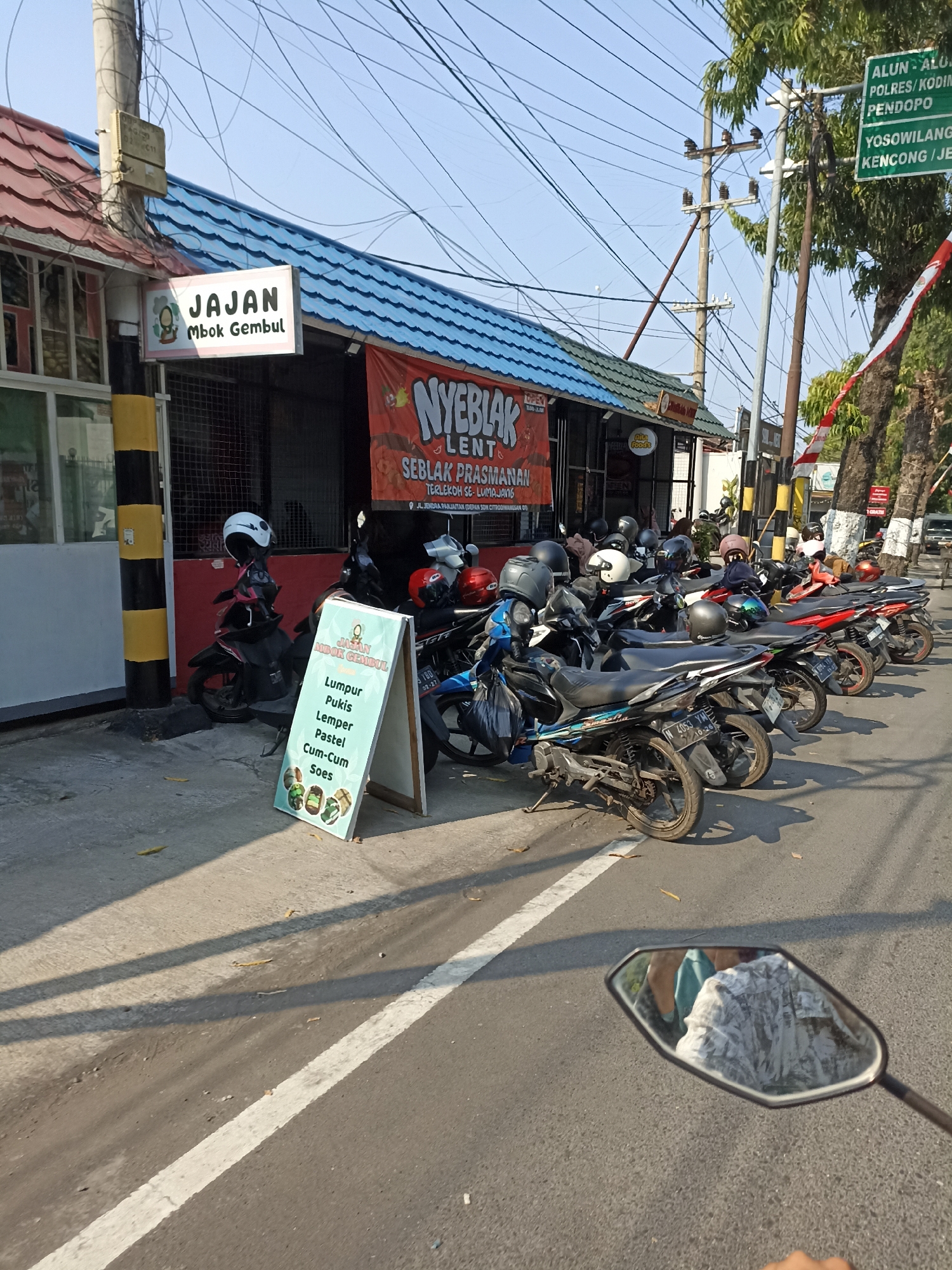In Citrodiwangsan, Indonesia, a street-side establishment named "NYEBLAK LENT" operates, selling food items such as "Lumpur Pukis," "Lemper," "Pastel," "Cum-Cum," and "Soes." The establishment is identified by a sign that reads "JAJAN Mbok Gembul." A row of motorcycles is parked in front of the establishment. A street signpost indicates directions to "ALUN - ALUN," "POLRES/KODIM," "PENDOPO," "YOSOWILANGUN," and "KENCONG / JEPARA." The scene is illuminated by sunlight, casting shadows on the asphalt road.